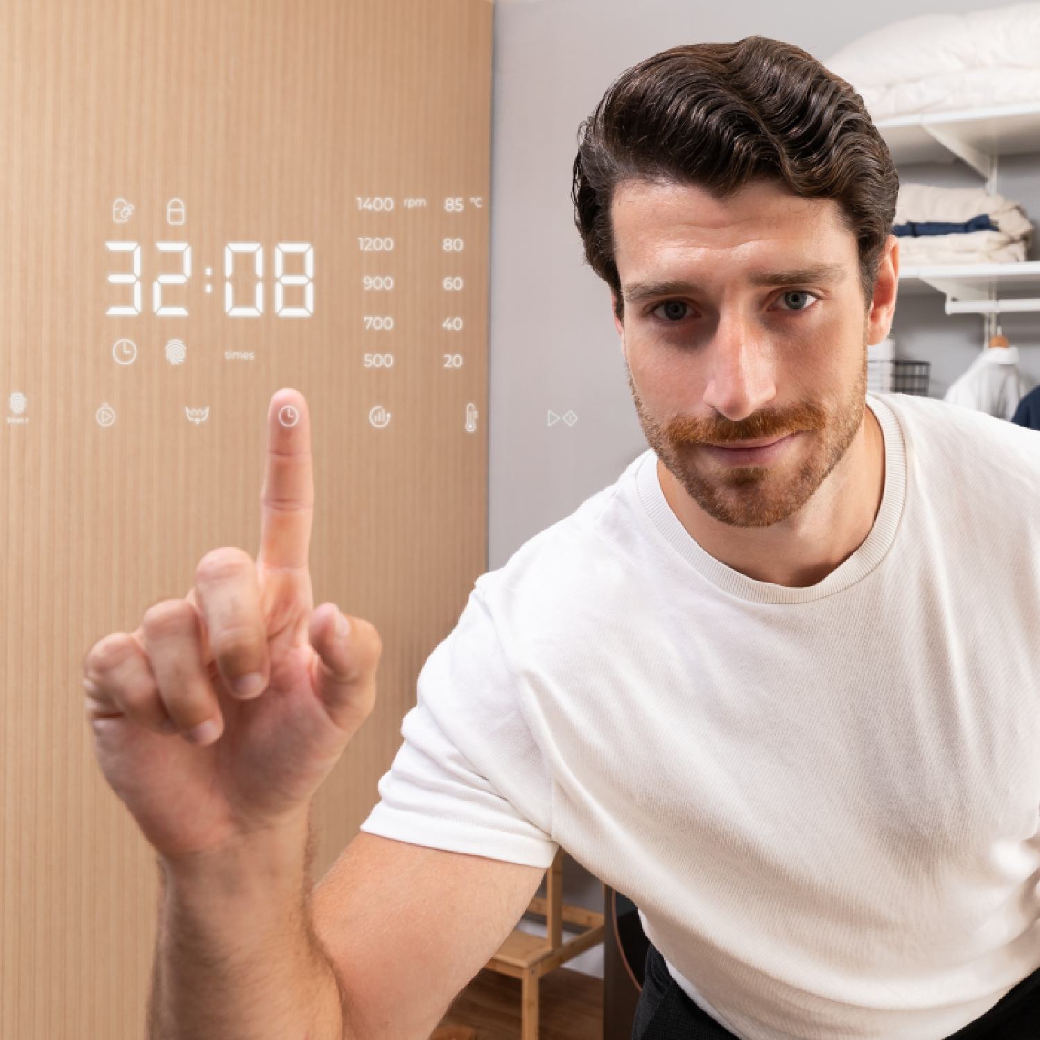 Un uomo indica un display digitale che mostra 32:08. Il display è su un pannello di legno. Indossa una maglietta bianca e guarda la fotocamera.