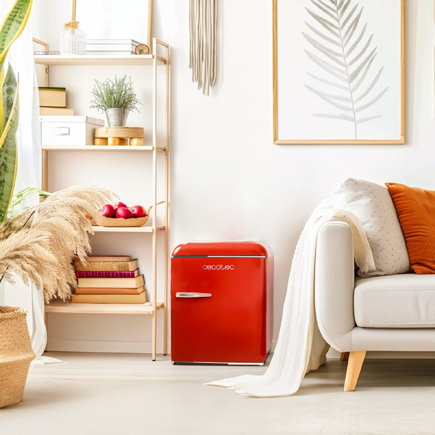 Un mini-frigo rosso accanto a un divano chiaro. Una mensola in legno con libri e piante. Opera d'arte con foglie incorniciata sul muro.