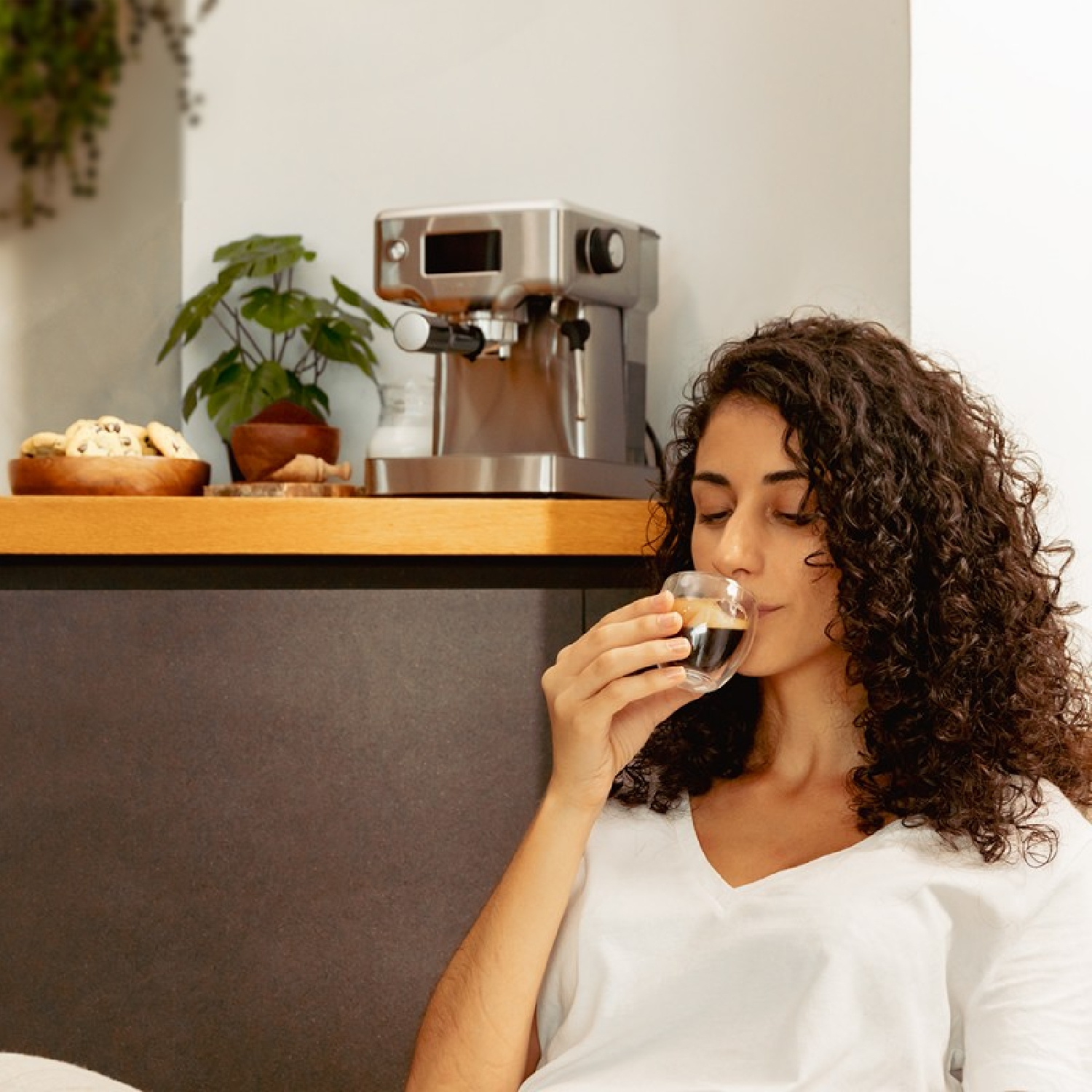 Kobieta z kręconymi włosami pije kawę ze szklanki. Za nią na blacie ekspres do kawy, ciasteczka i roślina.