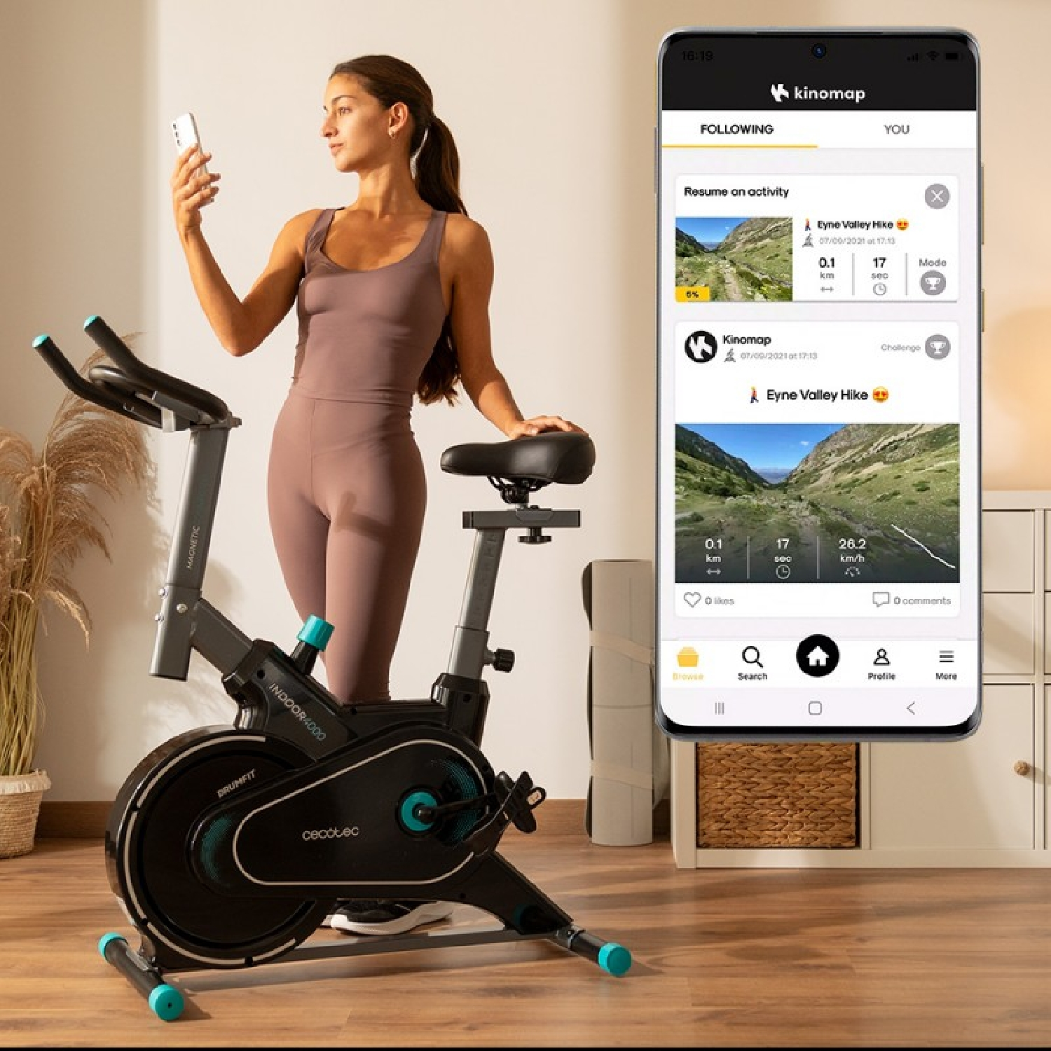 Mujer en bicicleta estática con teléfono, pantalla de la app visible. Fondo con cesta y esterilla de yoga.