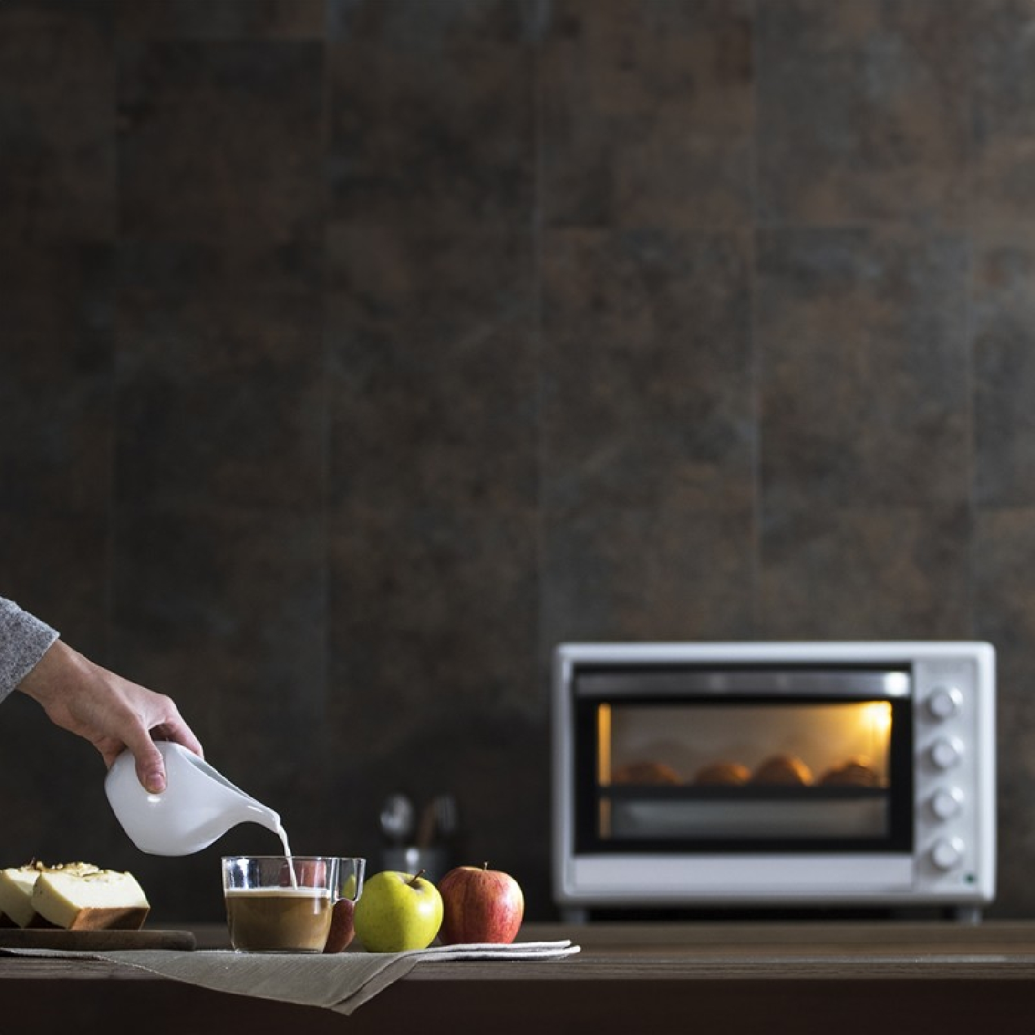 Una mano versa latte nel caffè. Mele, torta e un forno tostapane sono su un tavolo. Lo sfondo è un muro di piastrelle marroni.