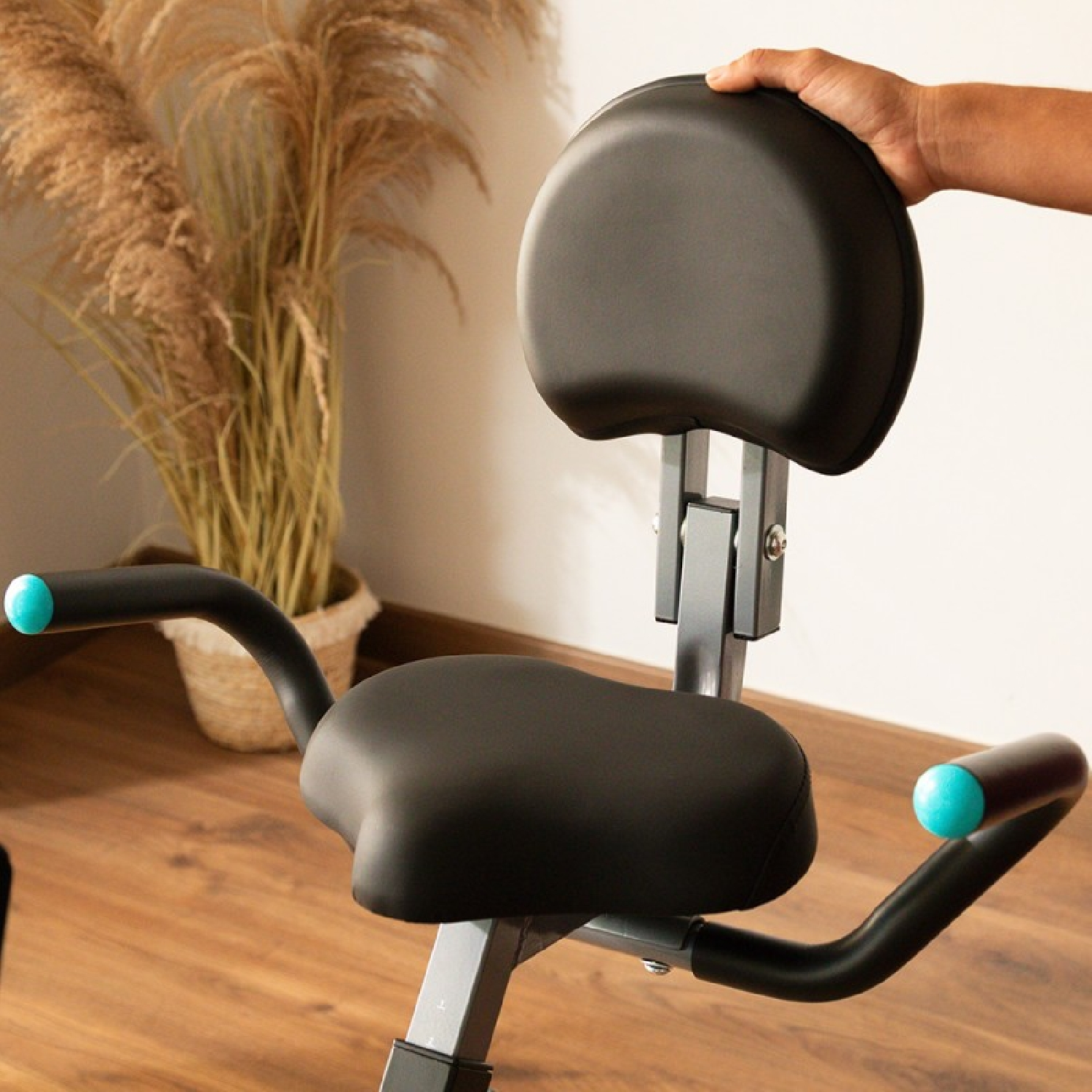 Dłoń na czarnym oparciu roweru stacjonarnego. Rower ma czarne siedzenie, podłokietniki z niebieskimi końcówkami i szarą ramę.