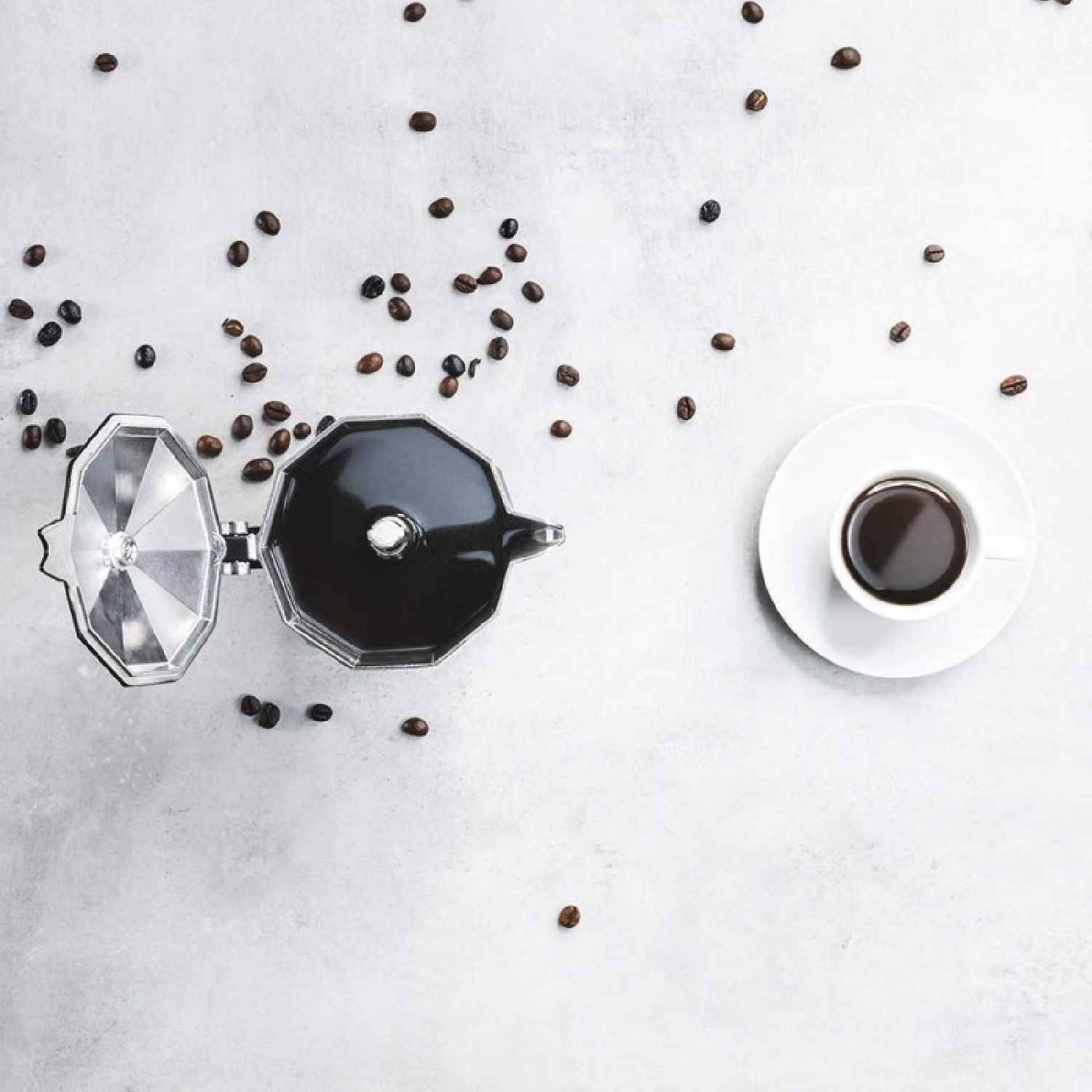 Vista dall'alto della preparazione del caffè. Chicchi di caffè sparsi attorno a una caffettiera moka e una tazza di caffè su una superficie bianca.