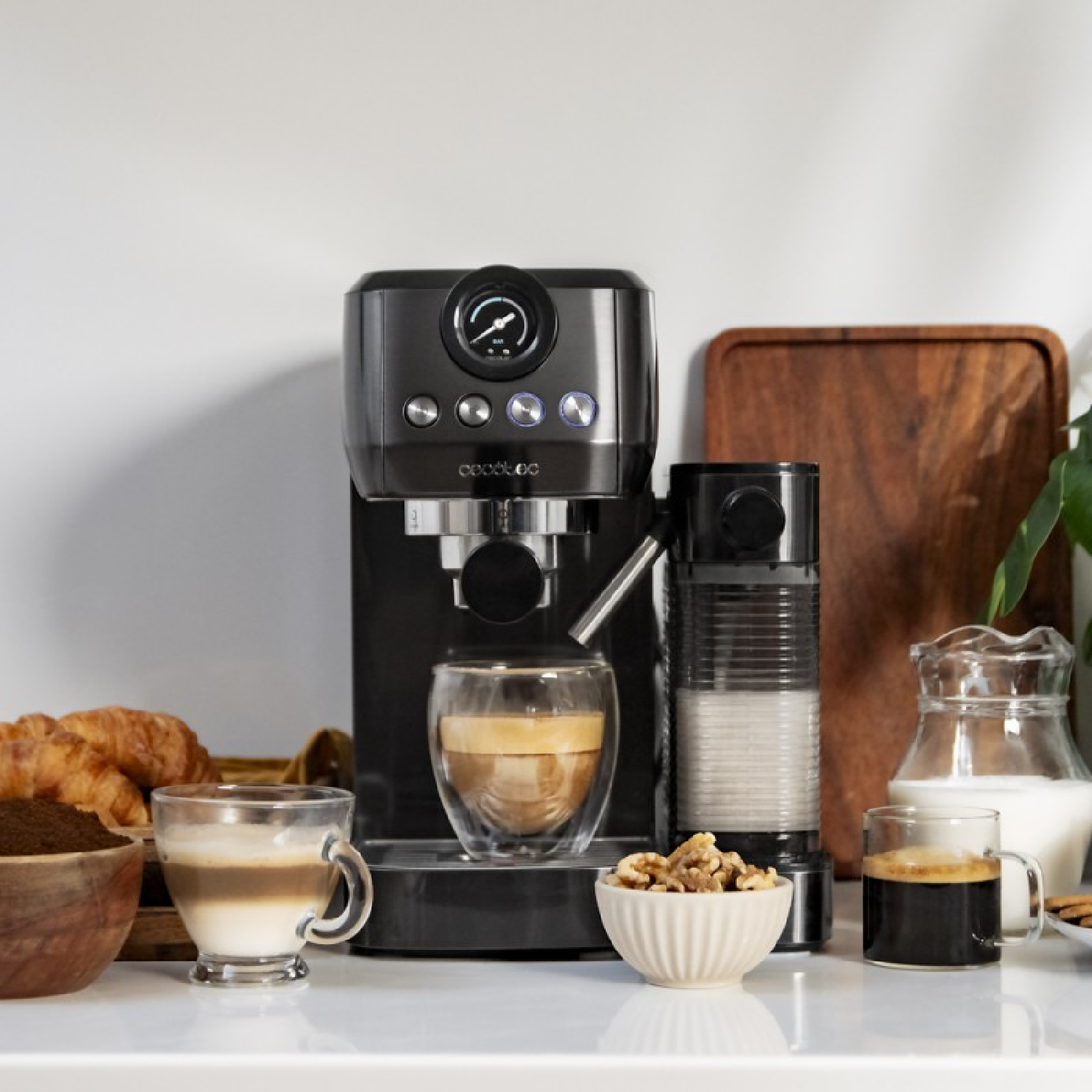 Una cafetera con café, leche, nueces y pasteles. La máquina es negra y plateada, y hay un vaso de café.