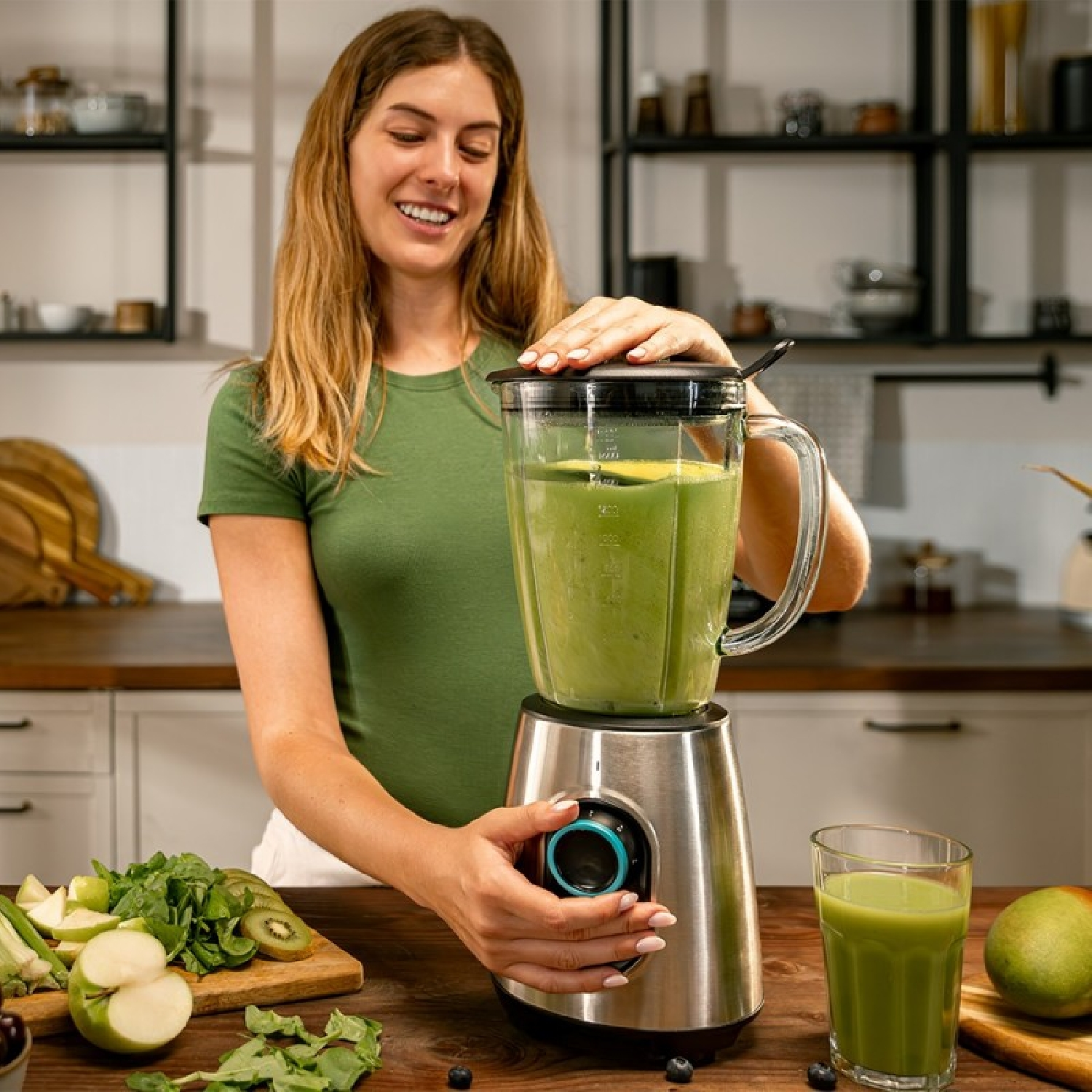Una donna prepara un frullato verde in un frullatore. Ingredienti come kiwi e mela sono sul tavolo. È presente un bicchiere di frullato.