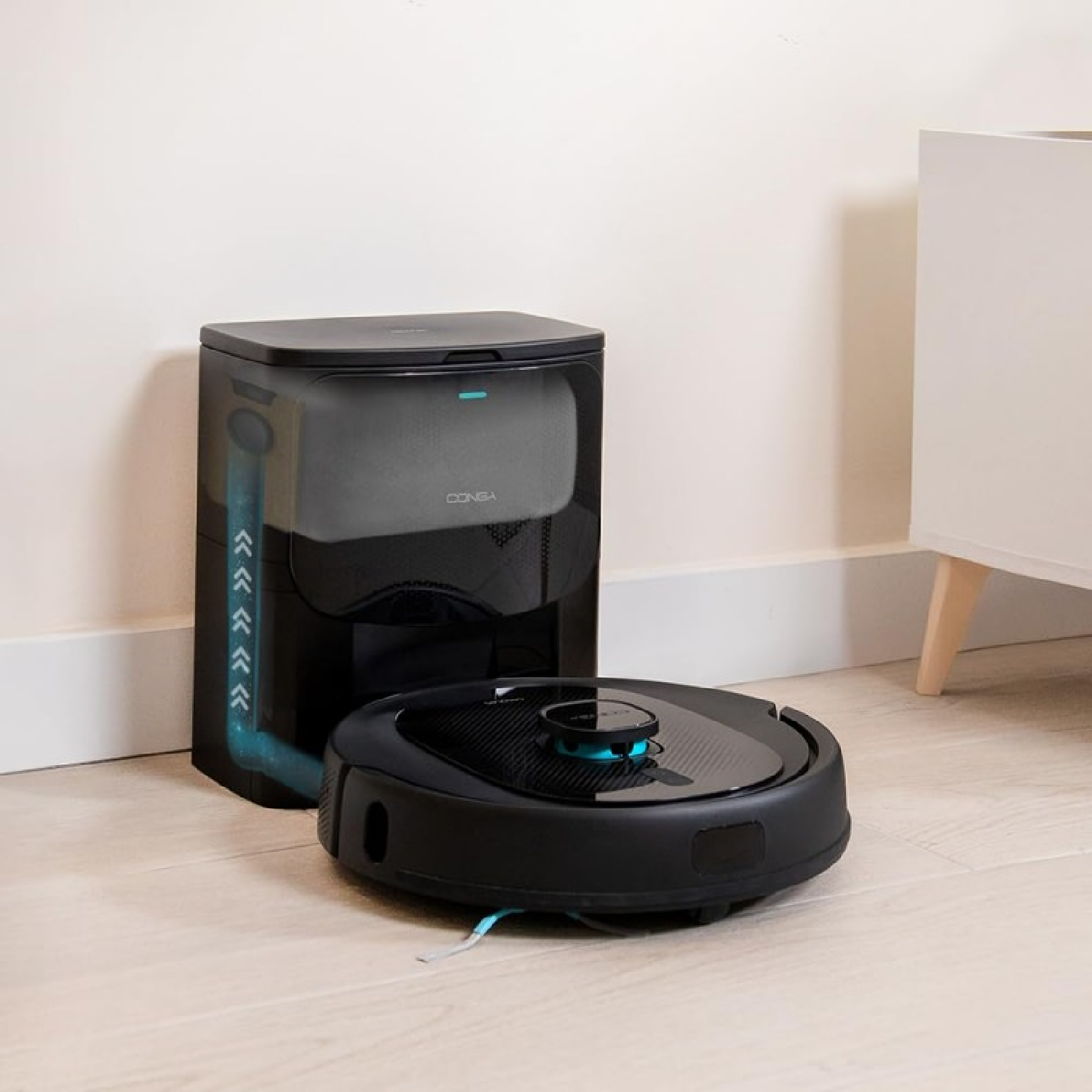 Un robot aspirapolvere nero e una stazione di ricarica si trovano su un pavimento in legno chiaro. La stazione è nera e ha un serbatoio d'acqua trasparente.