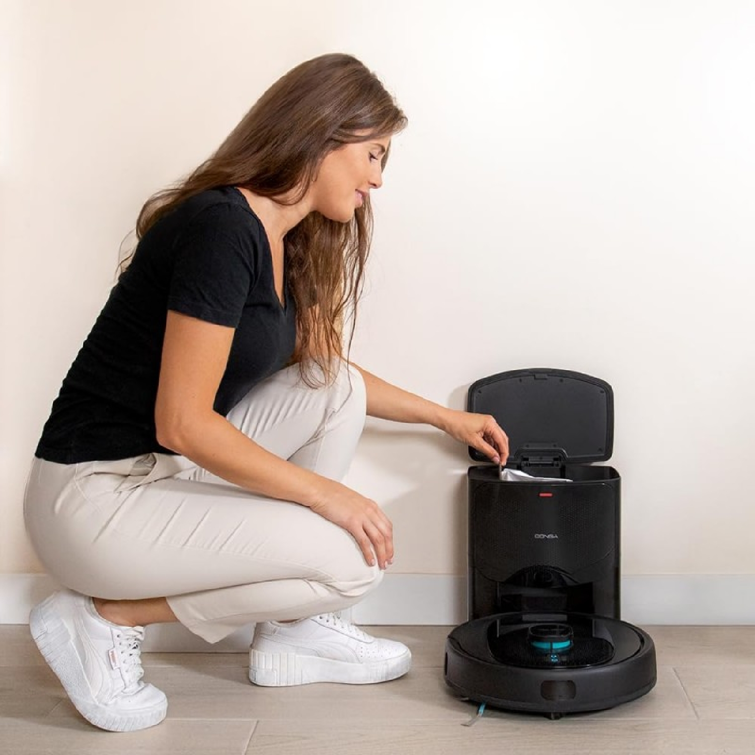 Una donna si accovaccia vicino a un robot aspirapolvere nero e alla sua stazione di ricarica, aprendo la parte superiore. Pareti e pavimento bianchi.