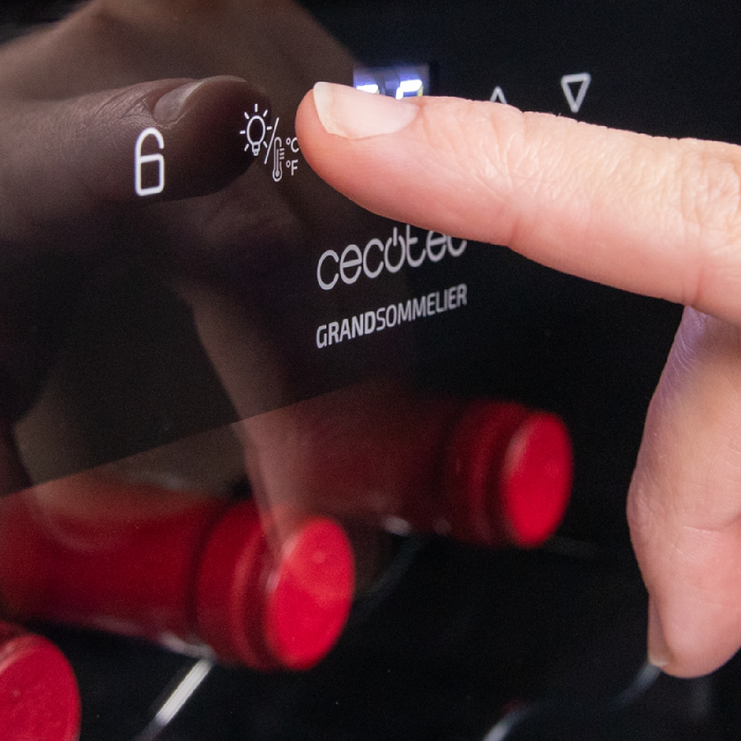 Un dito tocca un pulsante di una cantinetta. Il display mostra '6' e simboli di luce/temperatura. Si riflettono bottiglie di vino rosso.