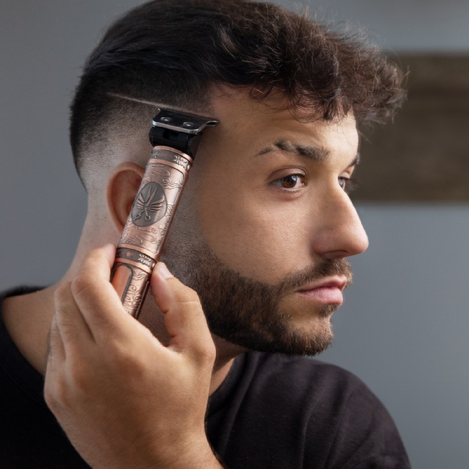 Un uomo si rade i capelli con un trimmer color rame. Ha occhi marroni, barba e capelli scuri. Lo sfondo è grigio.