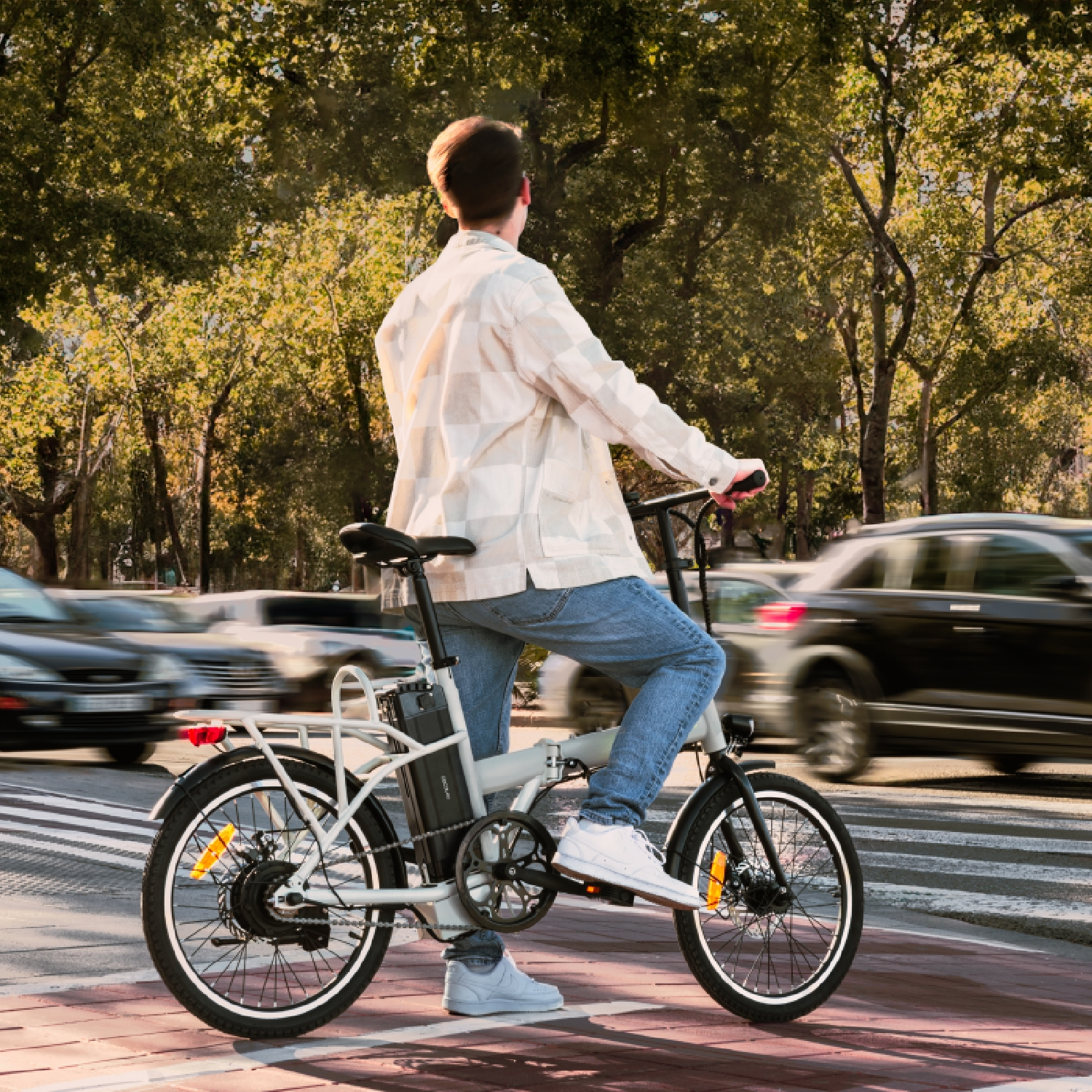 Una persona in sella a una e-bike su una pista ciclabile rossa con auto sullo sfondo. La persona indossa jeans e una camicia a quadri.