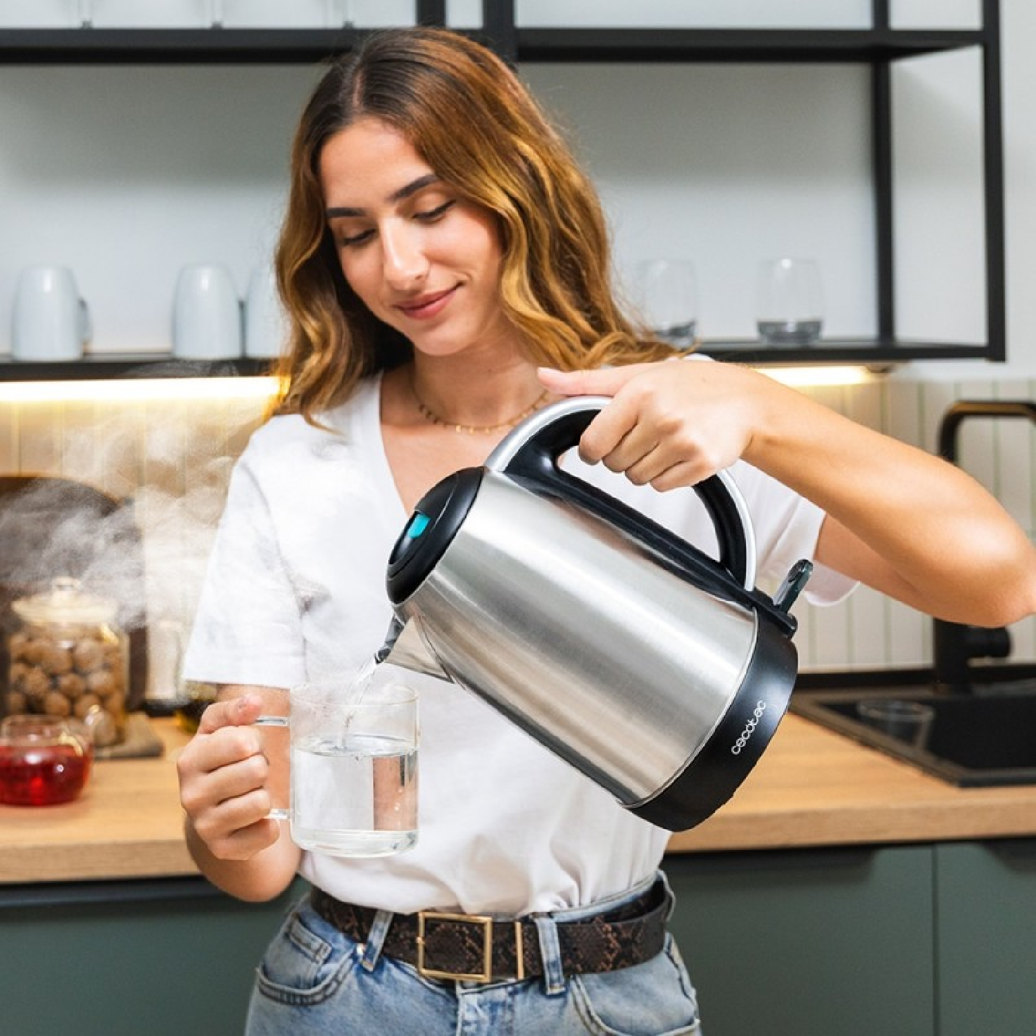 Una donna versa acqua calda da un bollitore in una tazza di vetro. Il vapore sale. Cucina sullo sfondo.