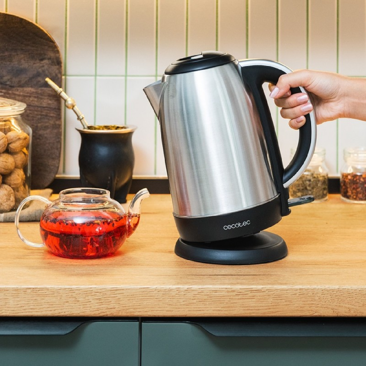 Una mano tiene un bollitore in acciaio. Accanto c'è una teiera di vetro con liquido rosso. Altri oggetti come barattoli e una tazza di mate sono visibili.