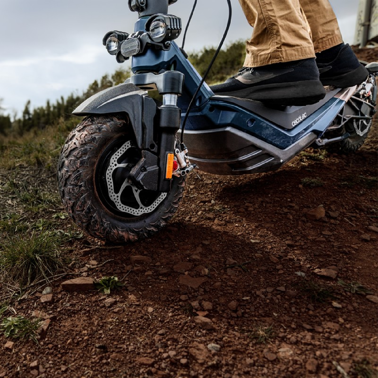 Una persona guida un monopattino elettrico blu su uno sterrato. Lo scooter ha pneumatici grandi. Il conducente indossa pantaloni marroni e scarpe nere.