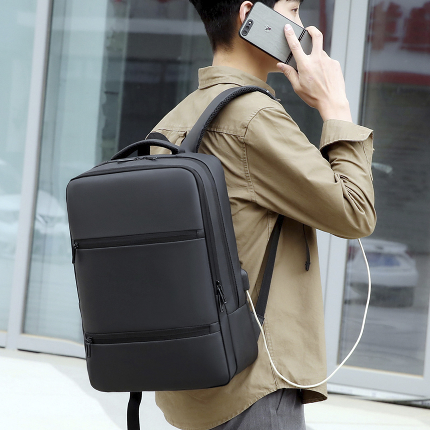 Un uomo con uno zaino nero parla al telefono. Indossa una giacca beige e pantaloni grigi. Lo sfondo è un edificio.