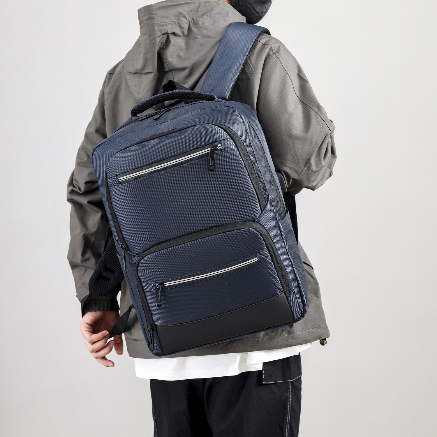 Una persona con una giacca grigia porta uno zaino blu navy con dettagli neri. Lo sfondo è bianco.
