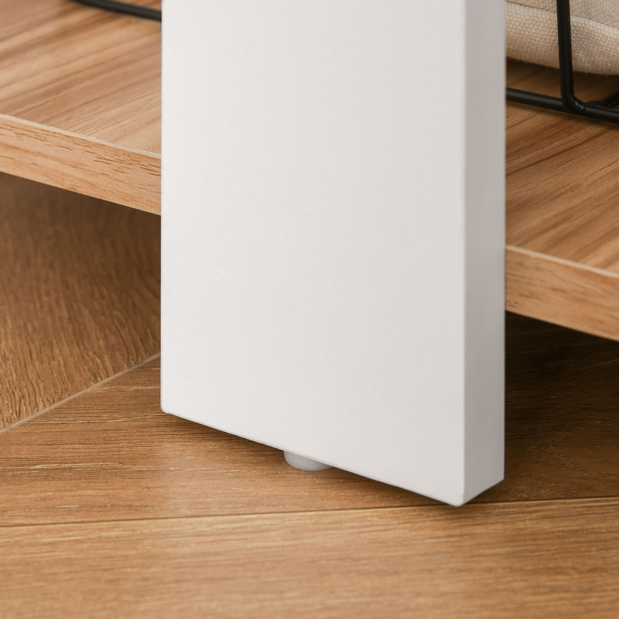 Primer plano de una pata de armario blanca sobre un suelo de madera. En el fondo se ven una estantería de madera y un marco de metal negro.