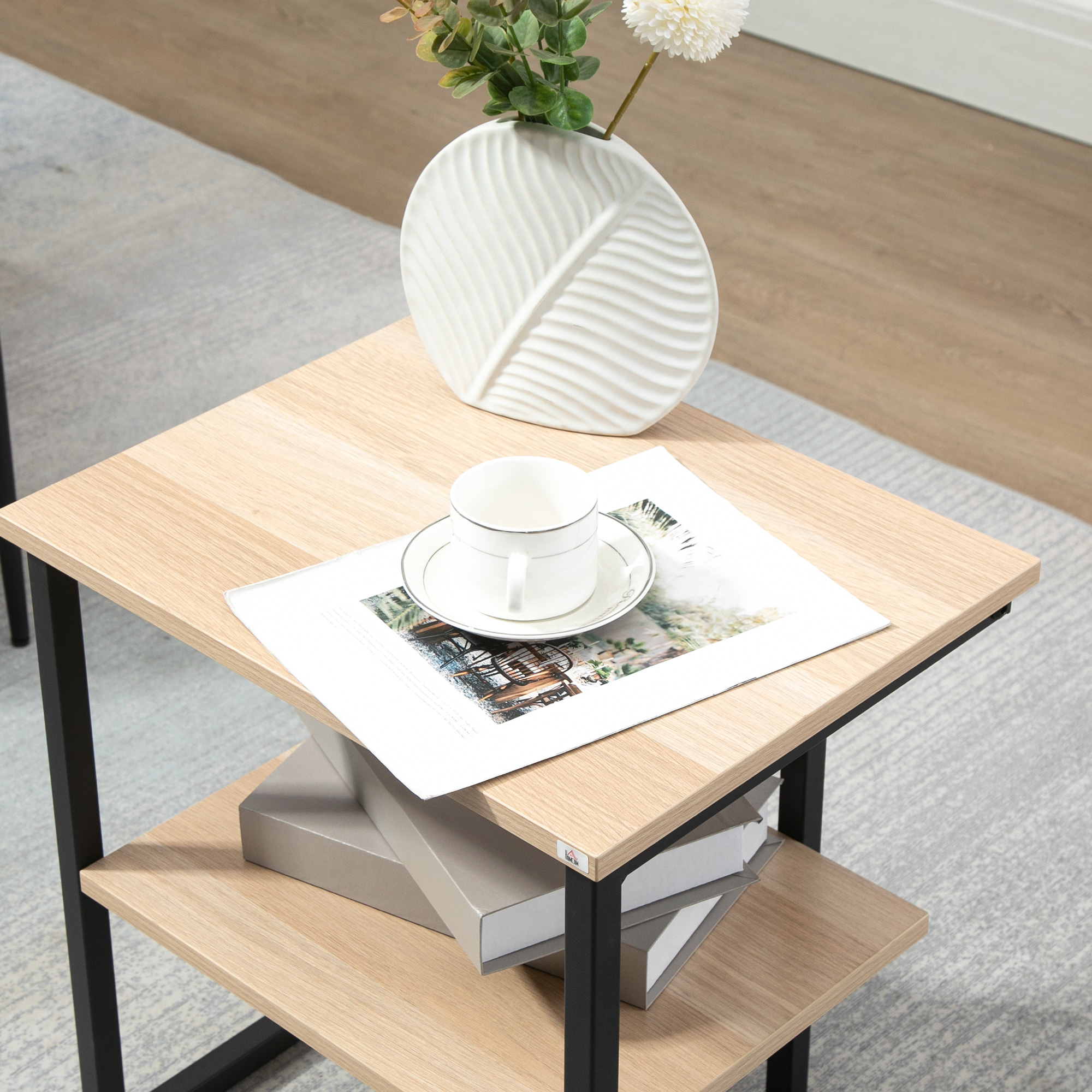 Una mesa cuadrada de madera con patas de metal negro. Un jarrón blanco con flores y una taza de té encima. Libros en el estante.