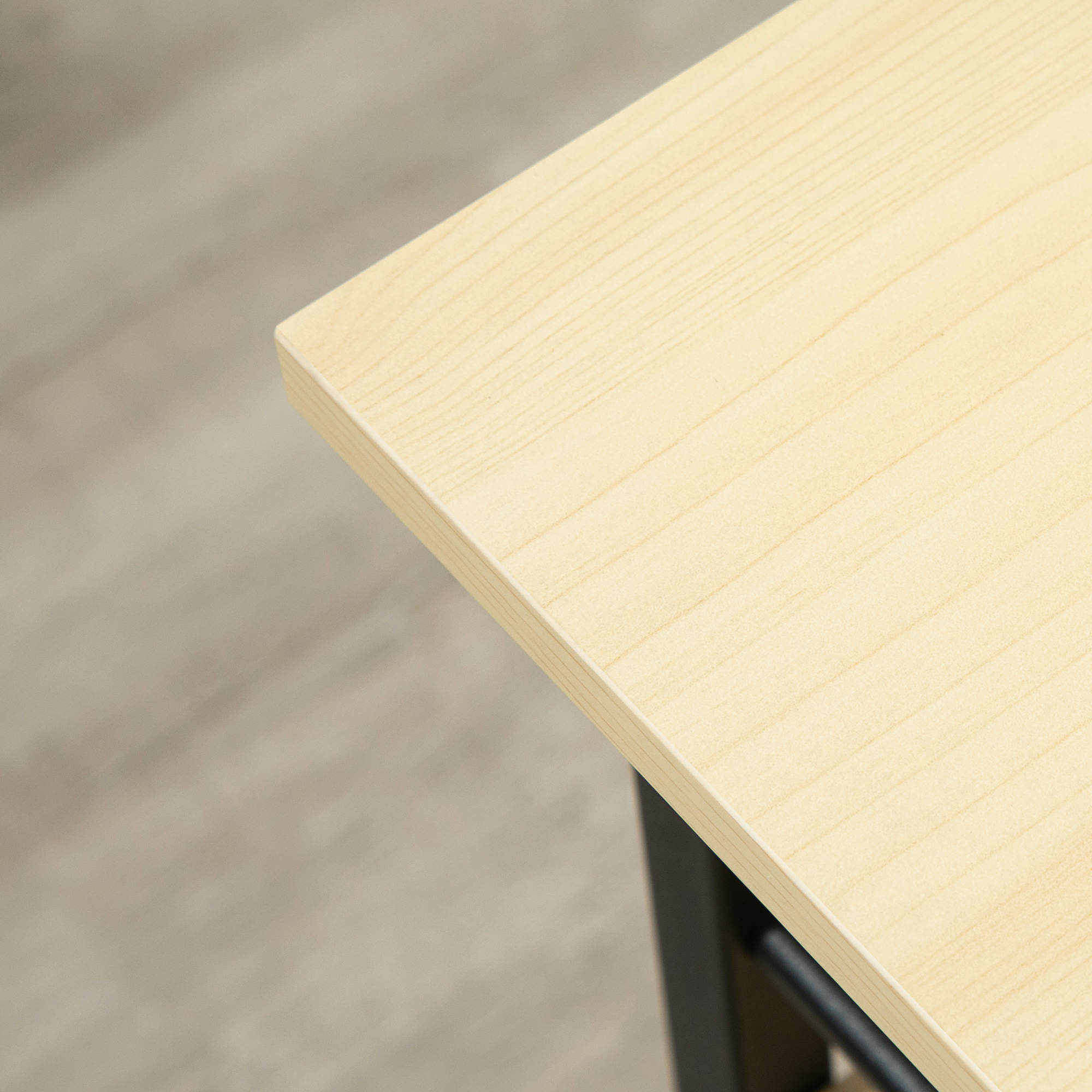 Primer plano de una esquina de mesa de madera clara con fondo borroso. Soporte de metal negro visible debajo de la mesa.