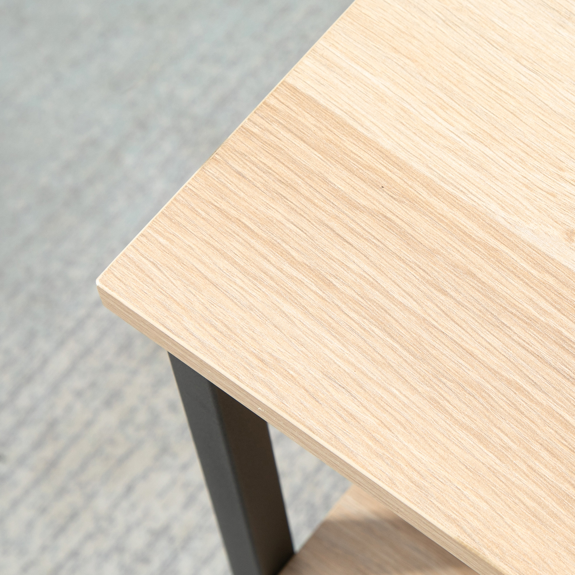 Primer plano de una esquina de mesa de madera. Veta de madera clara, pata negra. Suelo gris al fondo.