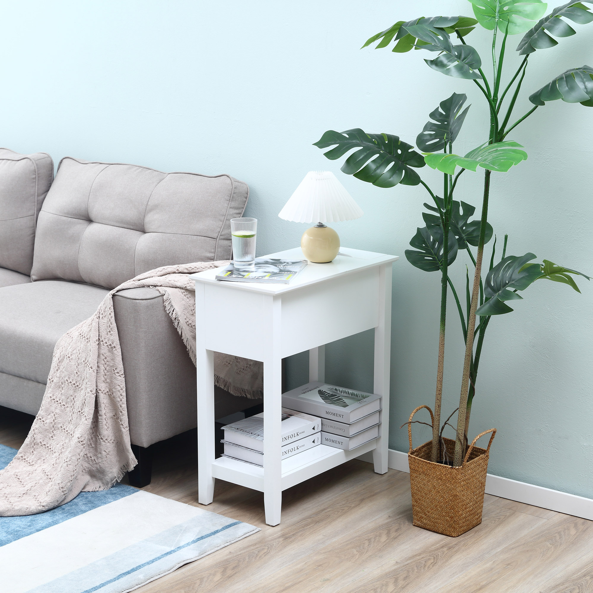 Una mesa auxiliar blanca con lámpara, libros y un vaso de agua junto a un sofá. Una planta grande a la derecha.