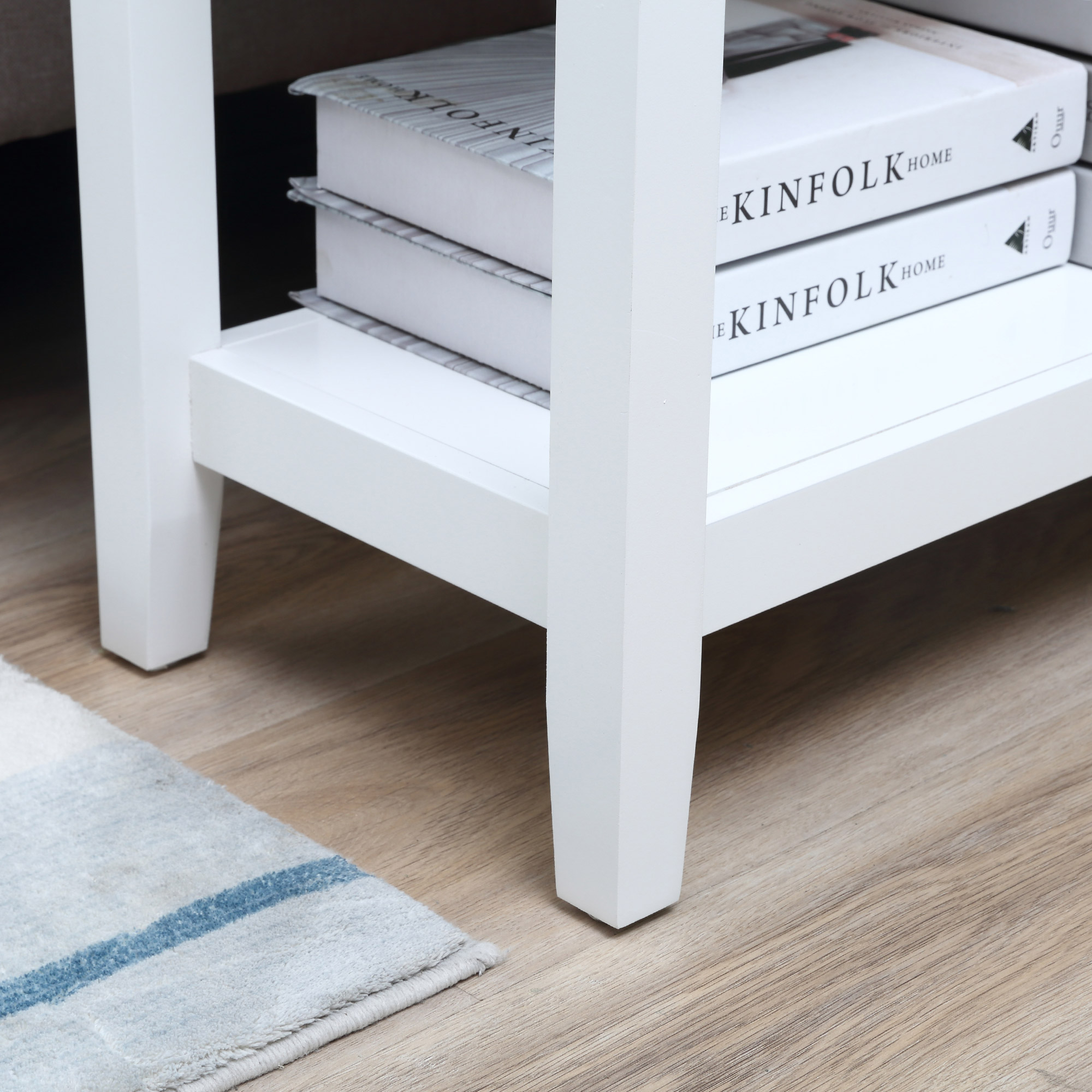 Mesa auxiliar blanca con libros, alfombra y suelo de madera. Los libros dicen 'Kinfolk Home'.