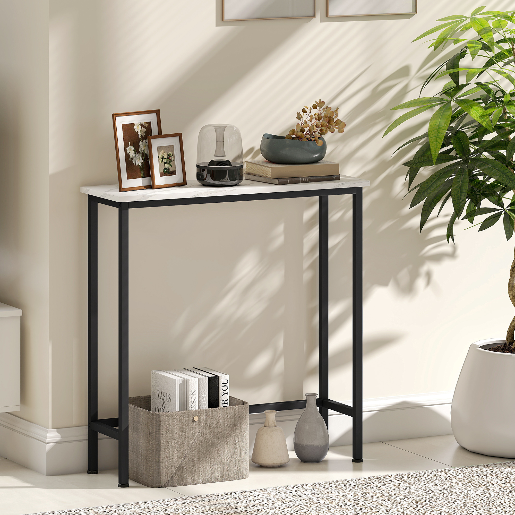 Una mesa con decoración: jarrones, libros y marcos. Hay una planta a la derecha. Sombras de persianas en la pared.