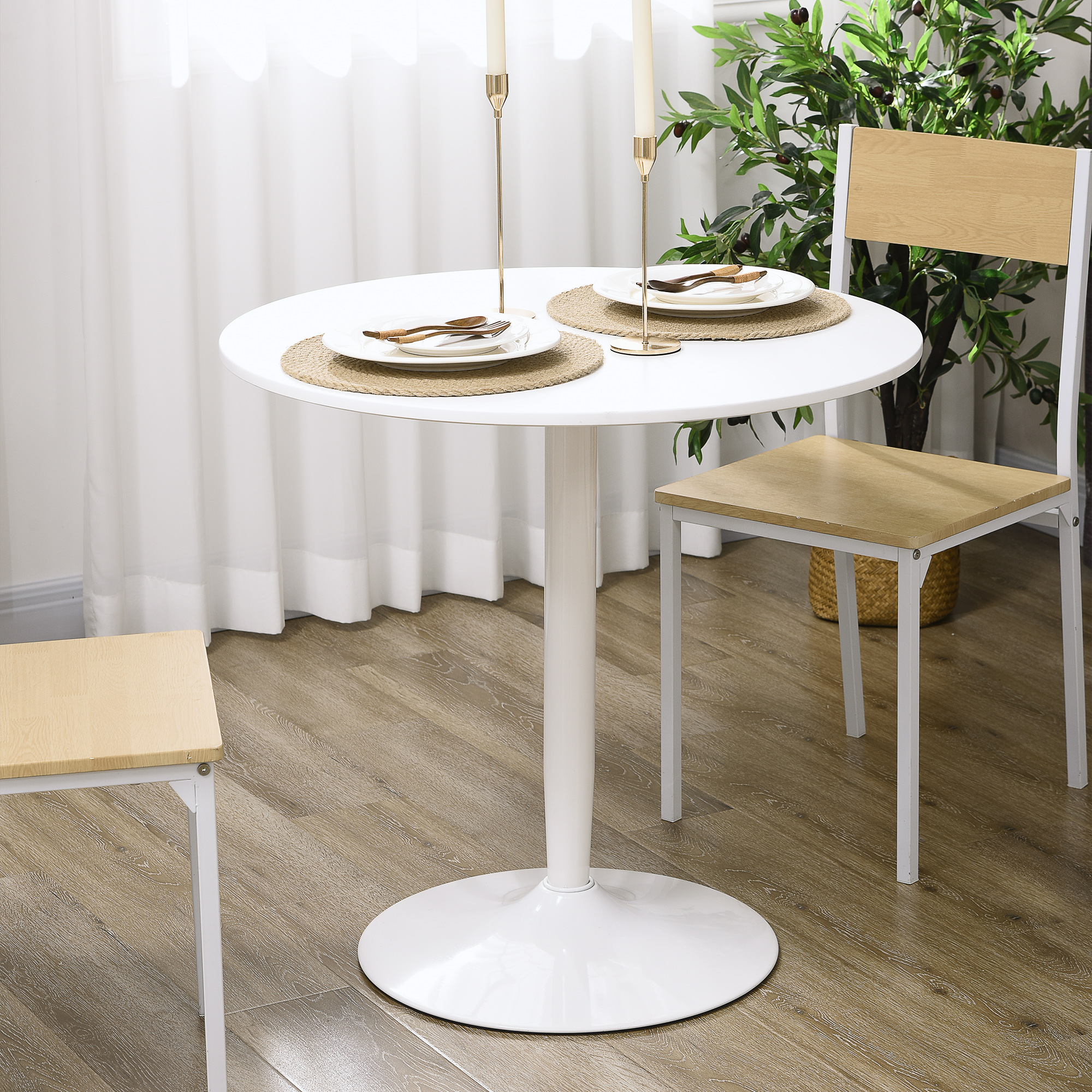 Una mesa redonda blanca con dos velas, dos sillas y platos. El fondo incluye una planta y una ventana con cortinas blancas.