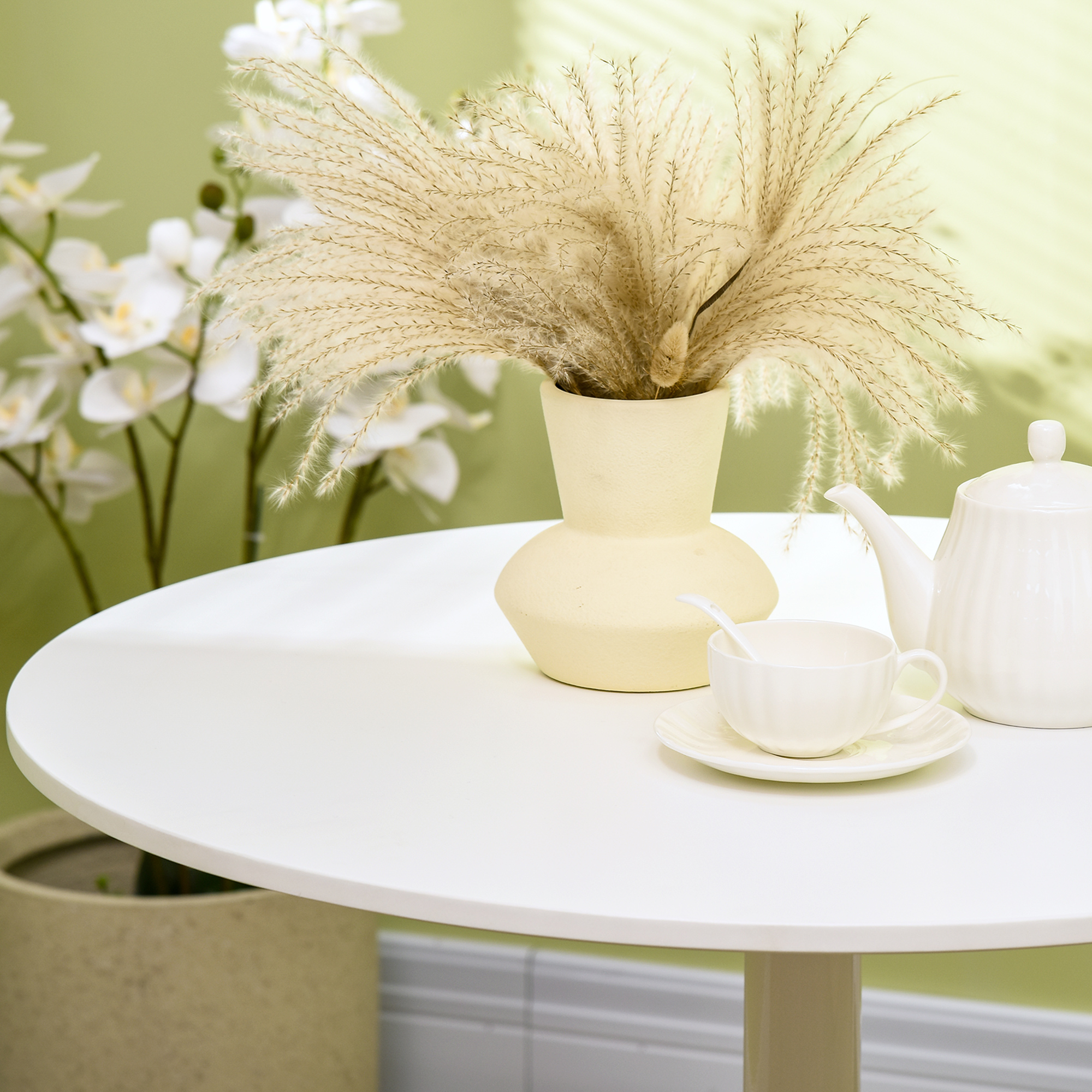 Una mesa blanca con taza, platillo, tetera y un jarrón con plantas secas. Fondo verde.