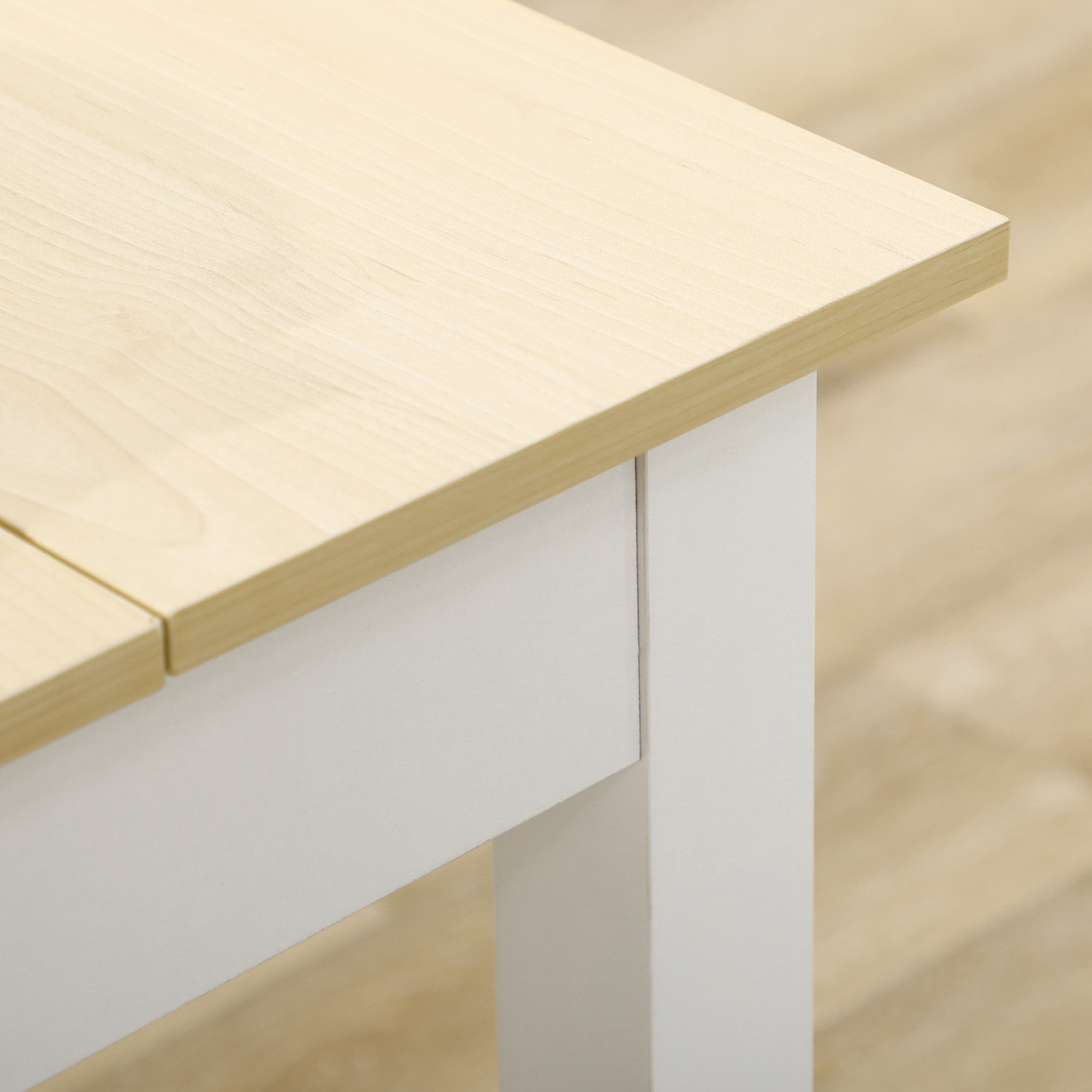 Primer plano de una esquina de mesa. La mesa es de madera clara, la base es blanca. Suelo de madera al fondo.