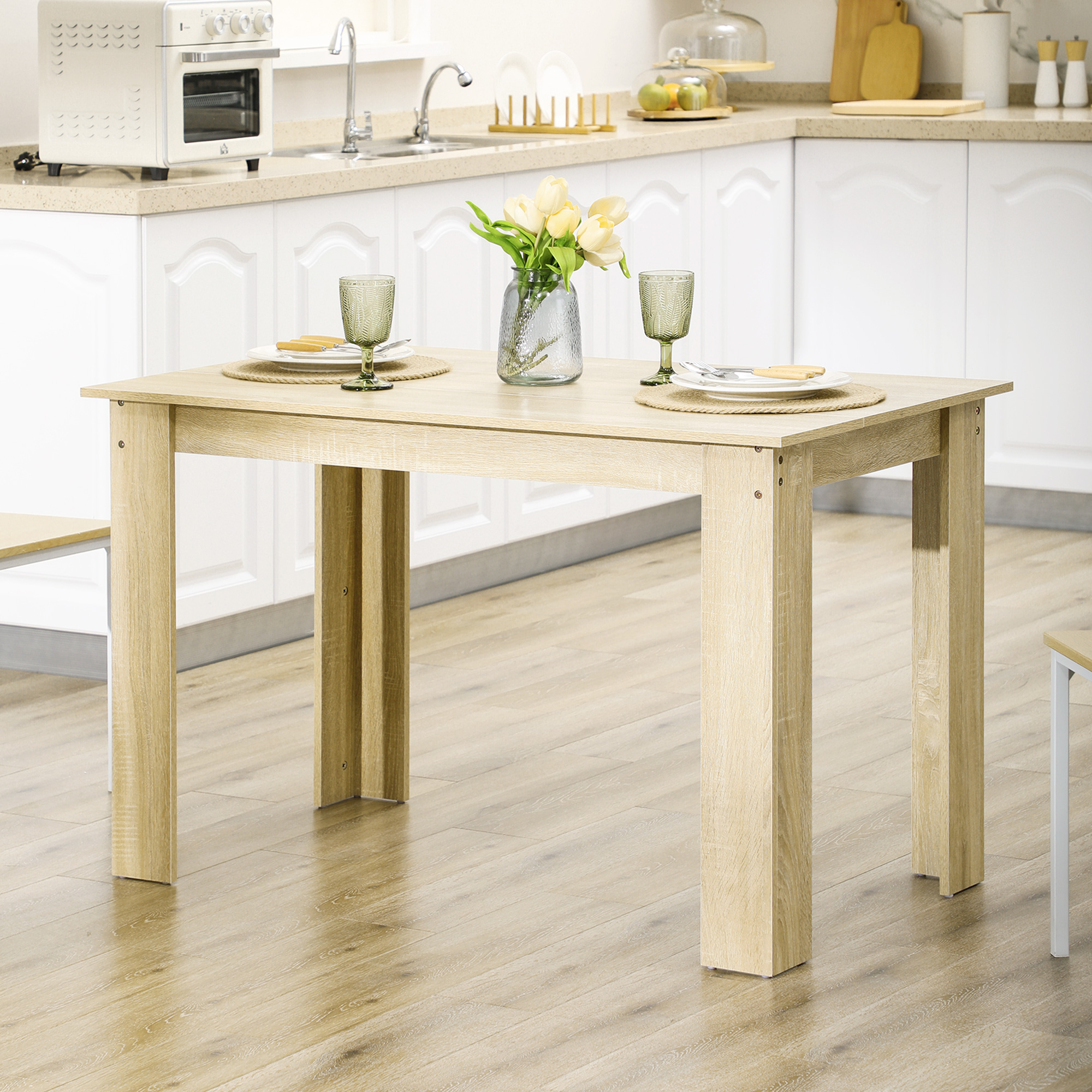 Una mesa de madera en un entorno de cocina. Tiene platos, vasos y un jarrón con flores. El fondo incluye armarios y electrodomésticos.