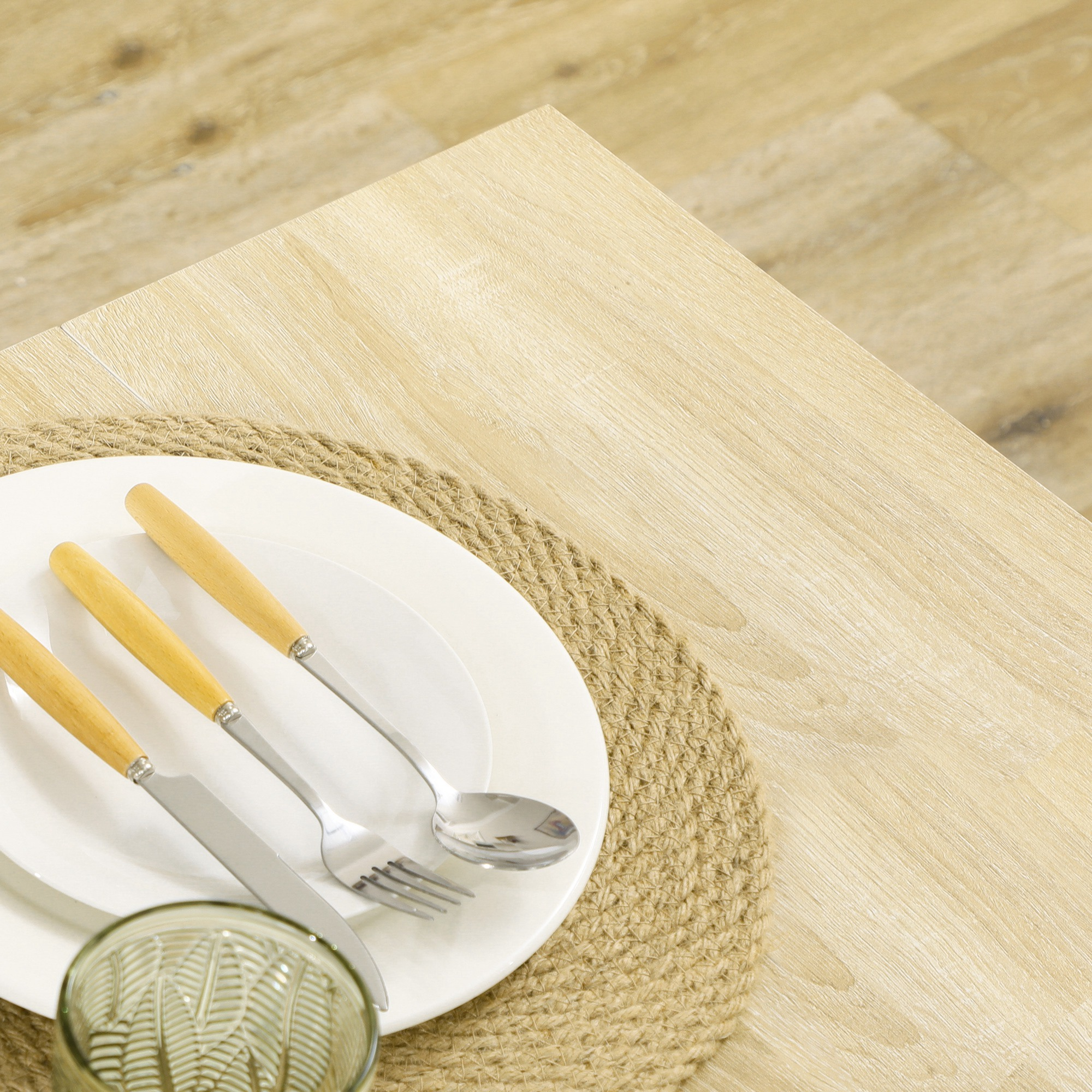 Servicio de mesa con platos blancos, cubiertos y un vaso. Se encuentra sobre un mantel individual tejido, sobre una mesa de madera.