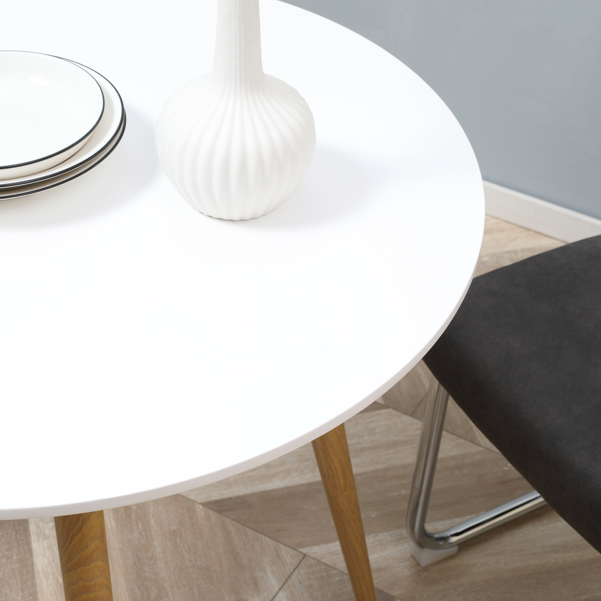 Una mesa blanca con platos, un jarrón y una silla. Suelo de madera. Pared azul claro al fondo.
