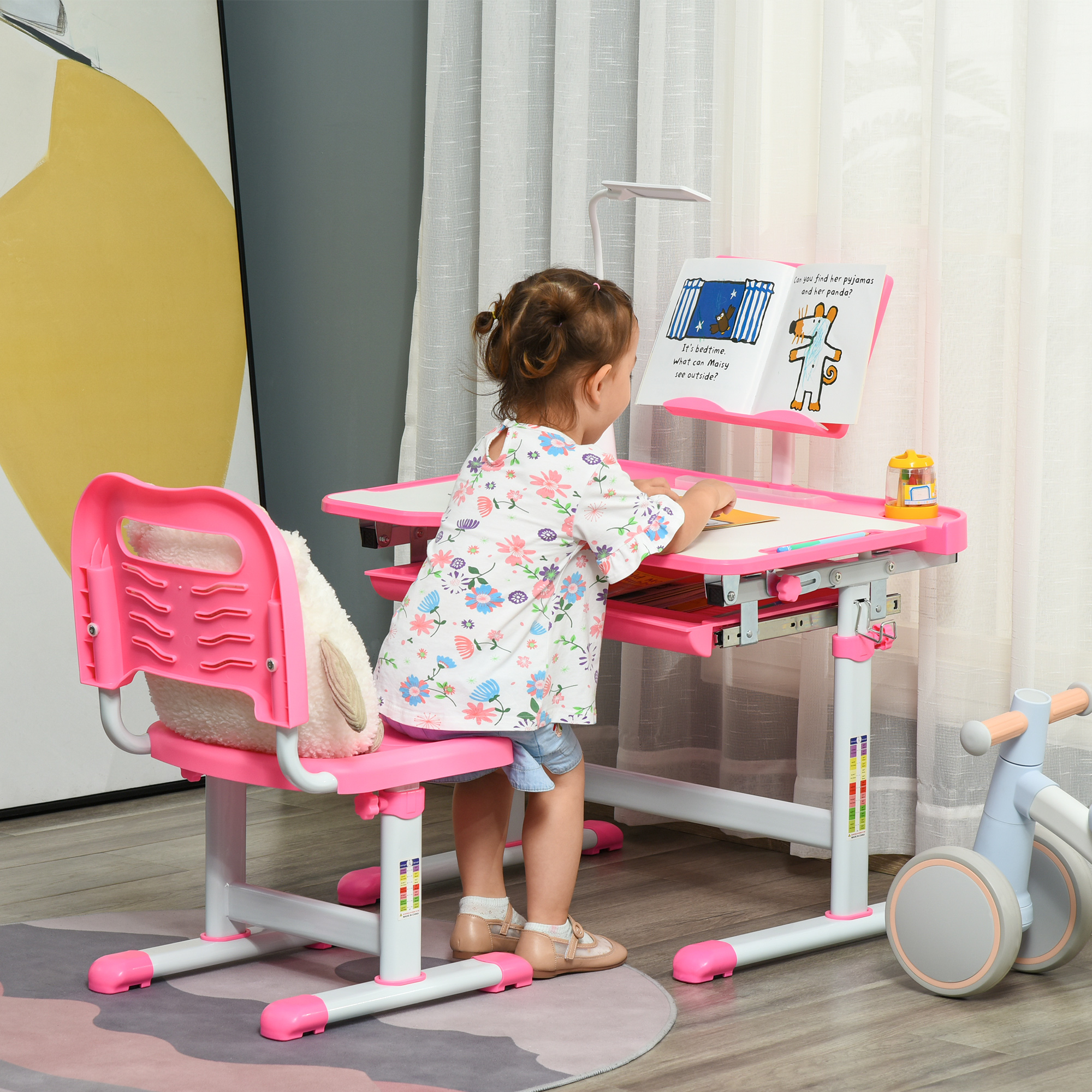 Una niña está en un escritorio rosa leyendo un libro. Un peluche está en la silla. Una bicicleta de juguete está en el fondo.