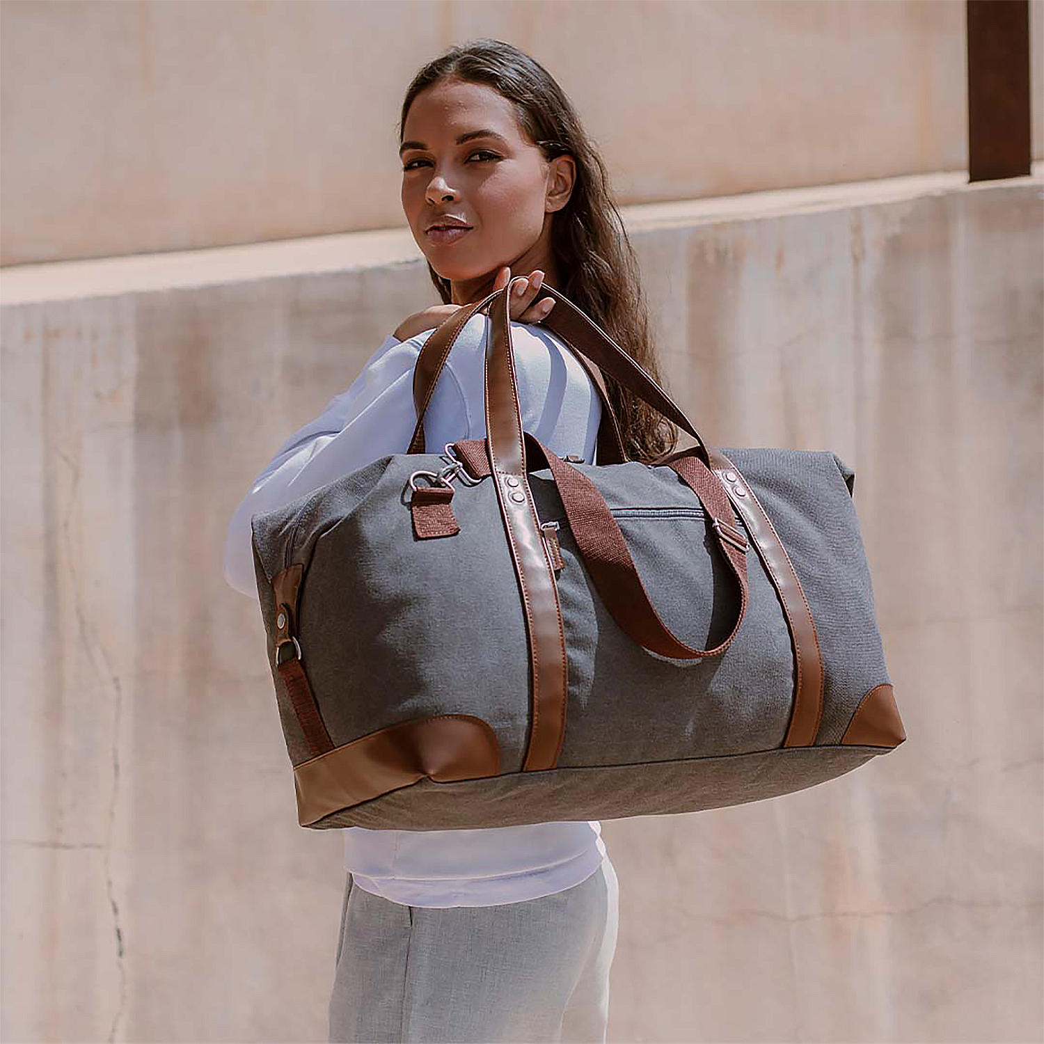 Una mujer sostiene una bolsa de lona gris con detalles en cuero marrón. Lleva camisa blanca y pantalón gris, con una pared beige detrás.
