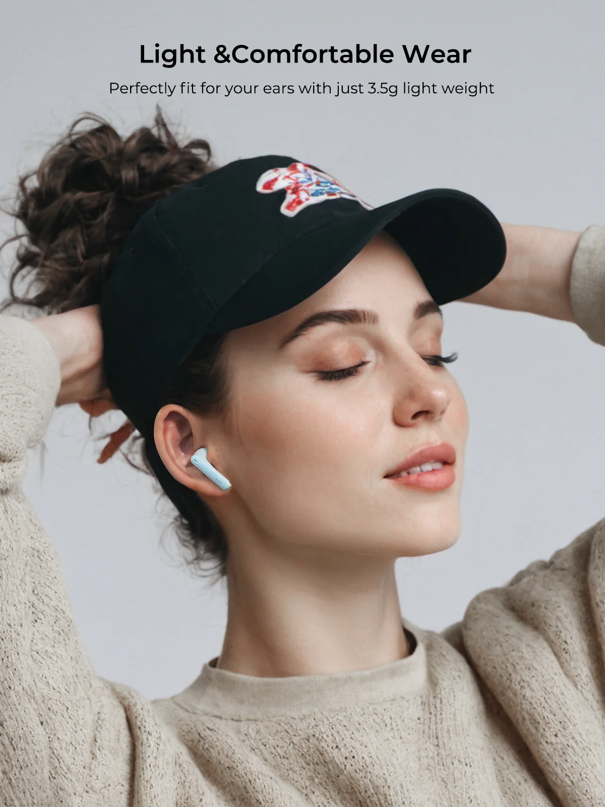 Una mujer lleva una gorra negra con un diseño y un auricular azul claro. Está sonriendo. Fondo blanco.