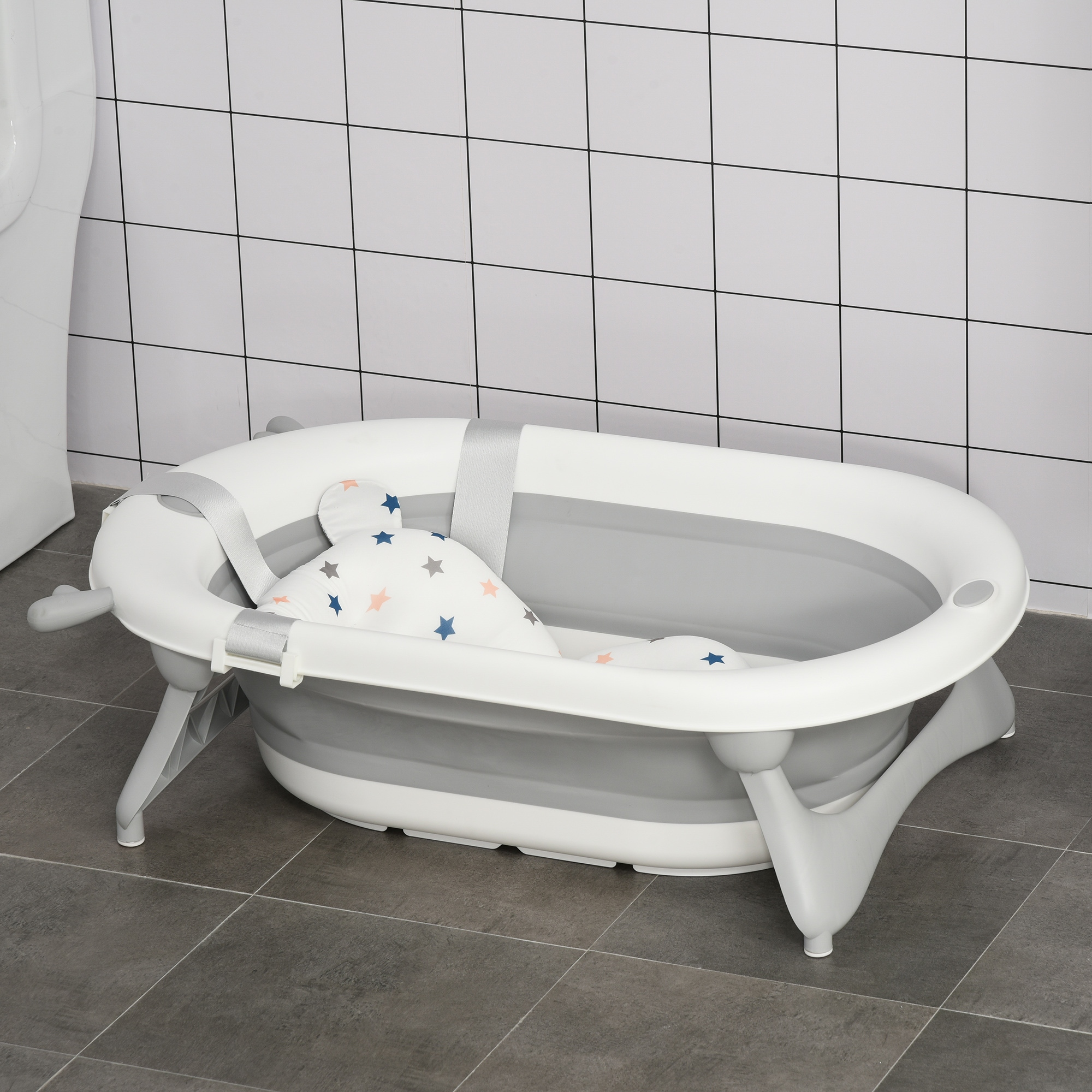 Una bañera para bebés blanca y gris con una almohada con estampado de estrellas en su interior. Está sobre un suelo embaldosado, contra una pared embaldosada.