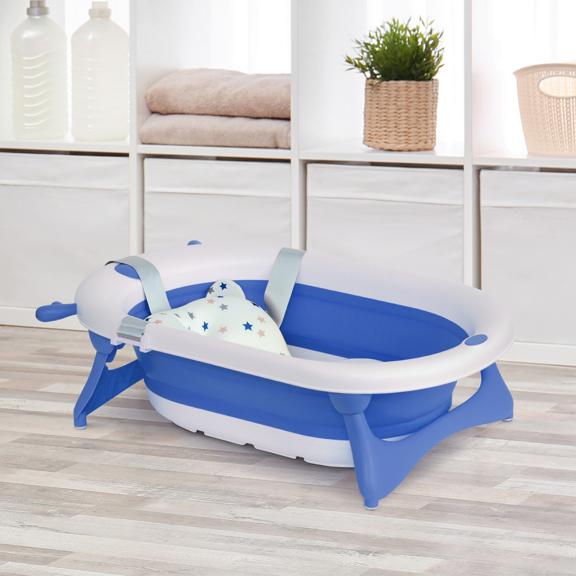 Una bañera para bebés azul y blanca con una almohada con estampado de estrellas en el interior. Estantes con toallas y plantas al fondo.