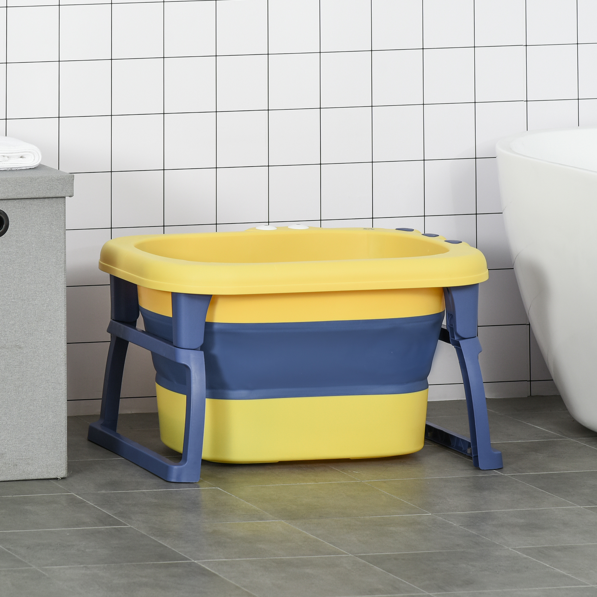Una bañera para bebés amarilla y azul con patas azules está colocada en un piso de baldosas grises, cerca de una bañera blanca y un armario gris.