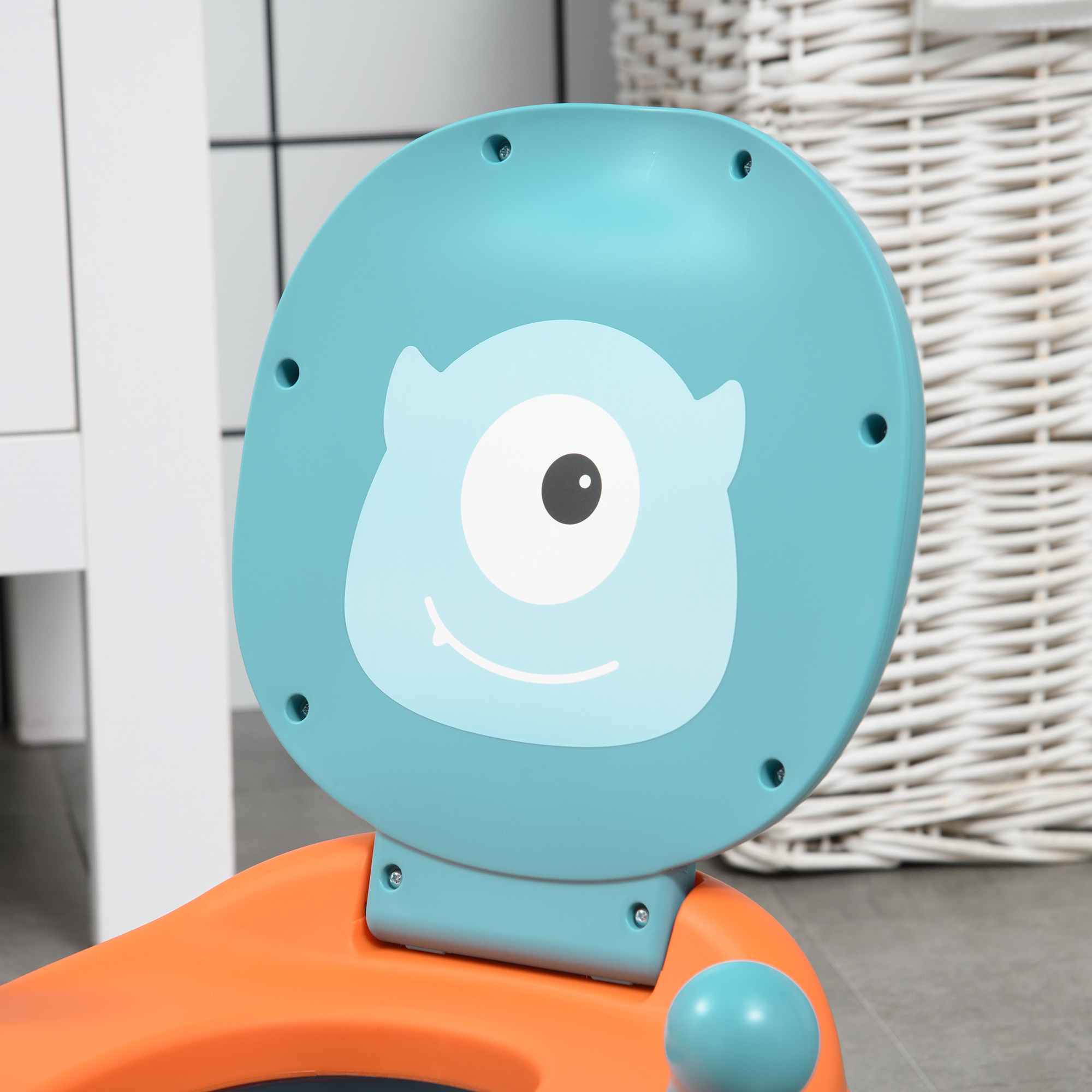 Un asiento azul para aprender a ir al baño con una cara de monstruo de dibujos animados está sobre una base naranja. Un cesto de mimbre blanco está detrás.