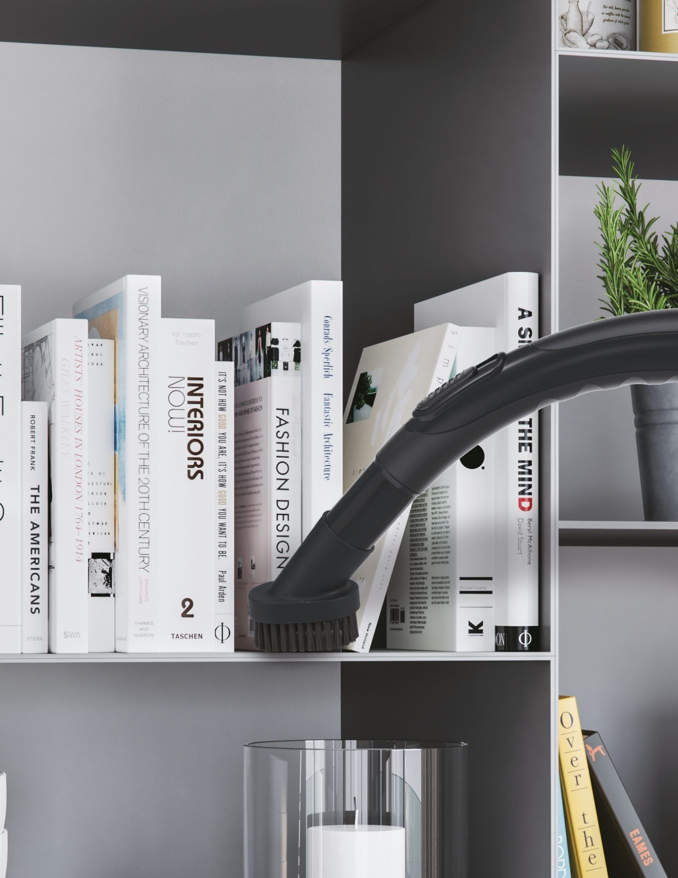 Odkurzacz czyszczący półkę z książkami. Na półce jest również roślina doniczkowa. Tło jest szare.