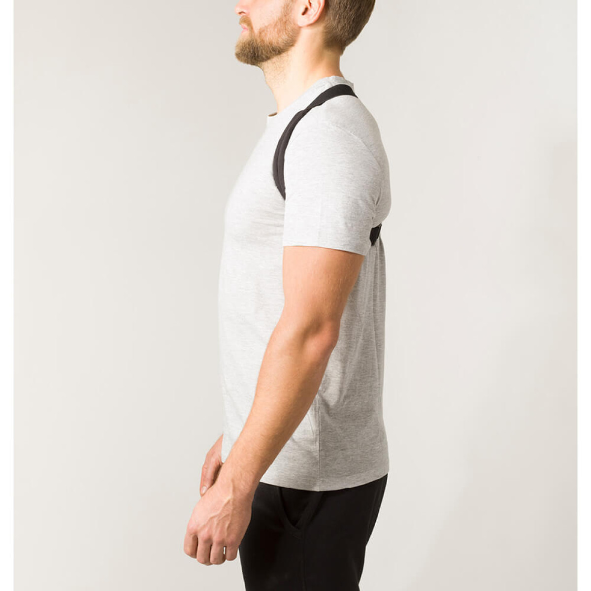 Ein Mann in grauem T-Shirt und schwarzer Hose, der eine Haltungskorrektur trägt. Er hat einen Bart und ist von der Seite zu sehen.
