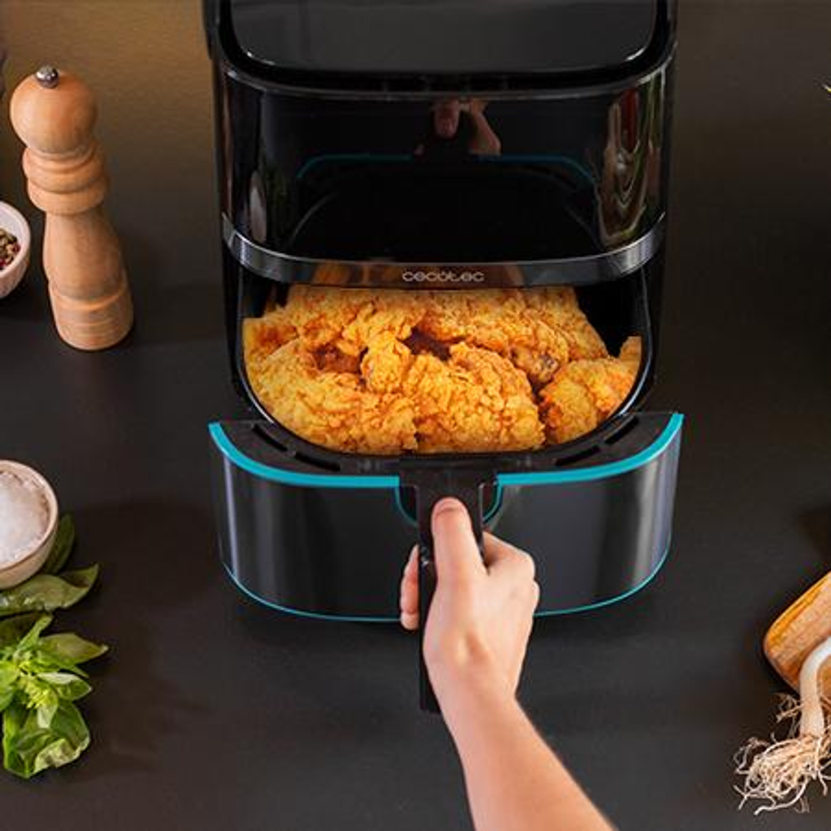 Una freidora de aire con pollo frito dentro es sostenida por una mano. Ingredientes como sal, pimienta y albahaca están al lado.