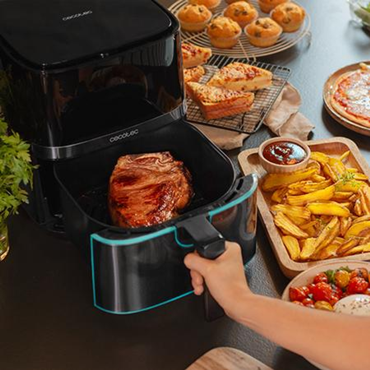 Una freidora de aire con un filete dentro, rodeada de comida cocinada como papas fritas, pizza, muffins y salsas en una mesa. Una mano sostiene la freidora.