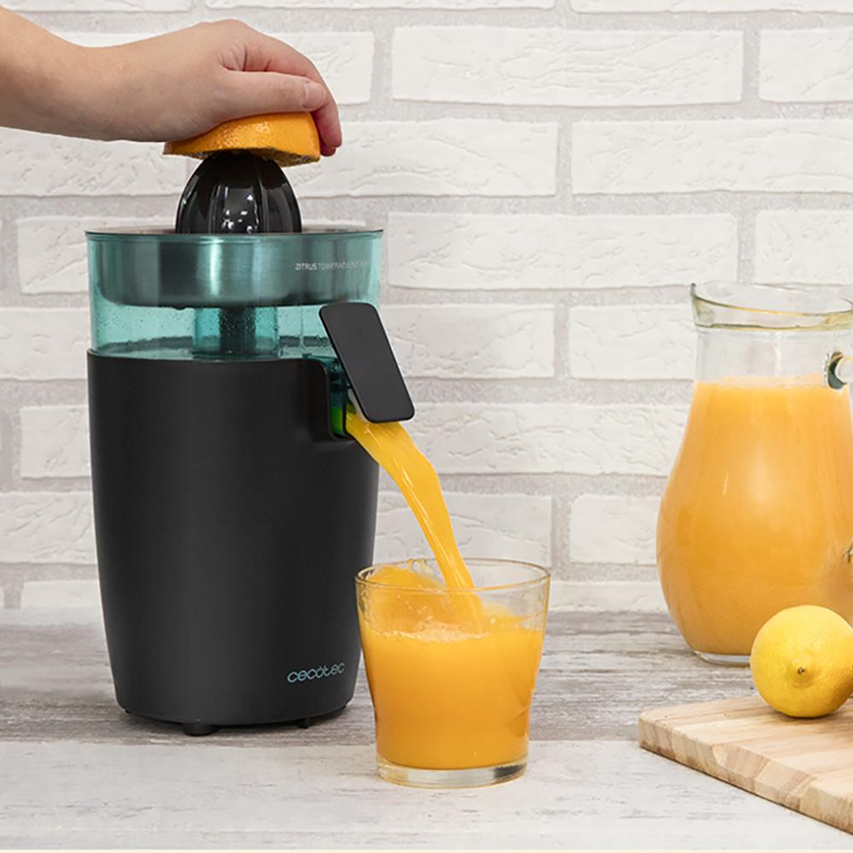 Una mano exprime media naranja en una exprimidora. El zumo fluye a un vaso. Una jarra y un limón están en una tabla de madera. Pared de ladrillos blancos.