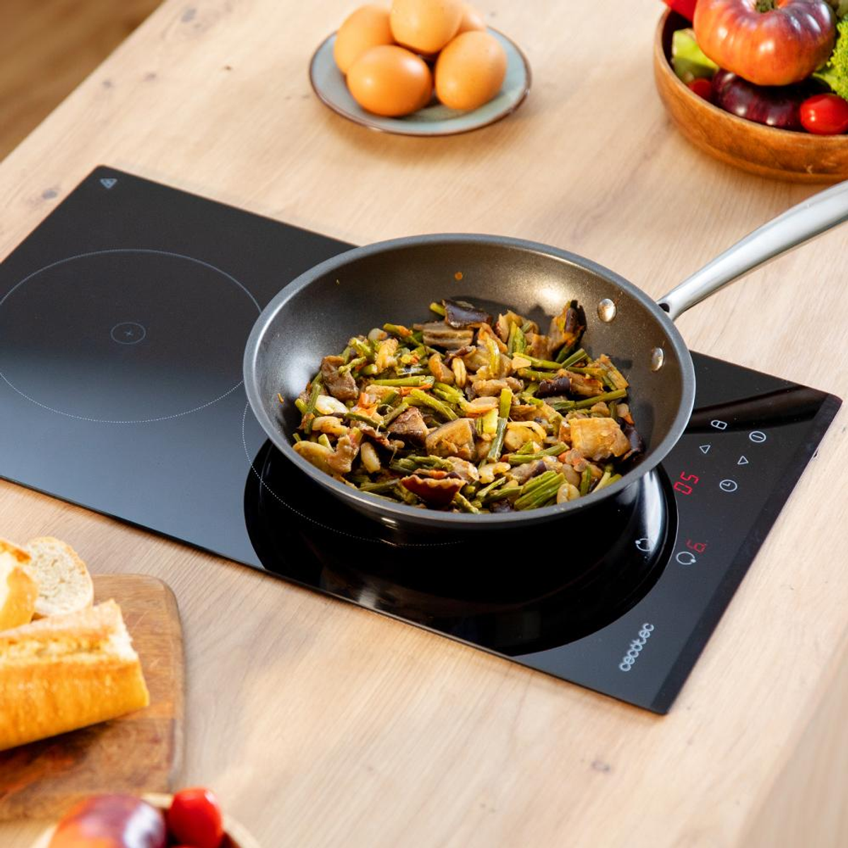 Una sartén con verduras cocinadas sobre una estufa negra. Huevos, pan y tomates están en la encimera de madera.