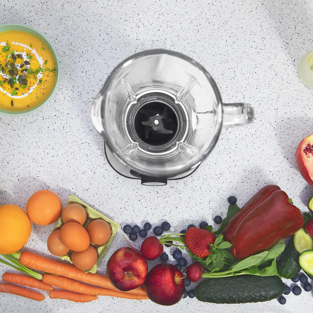 Una licuadora rodeada de frutas y verduras. Incluye naranjas, zanahorias, huevos, arándanos, manzanas, pimientos y pepinos.