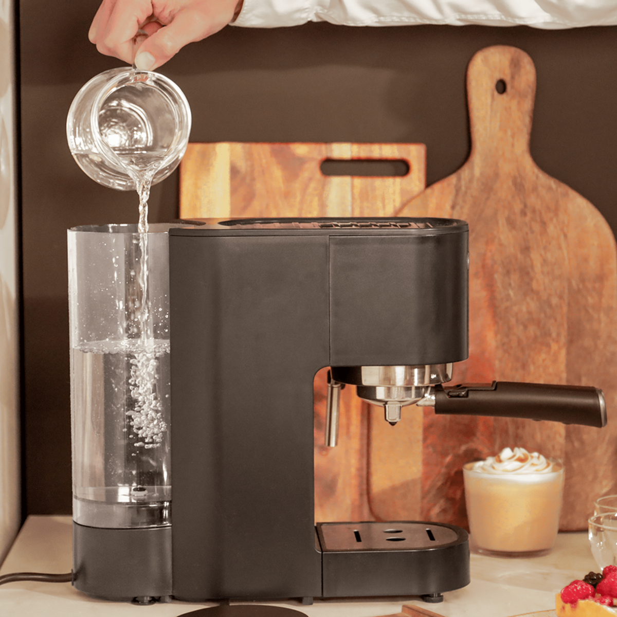 Eine Person gießt Wasser aus einer Glaskaraffe in eine schwarze Espressomaschine. Holzschneidebretter und ein Kaffeegetränk sind im Hintergrund.