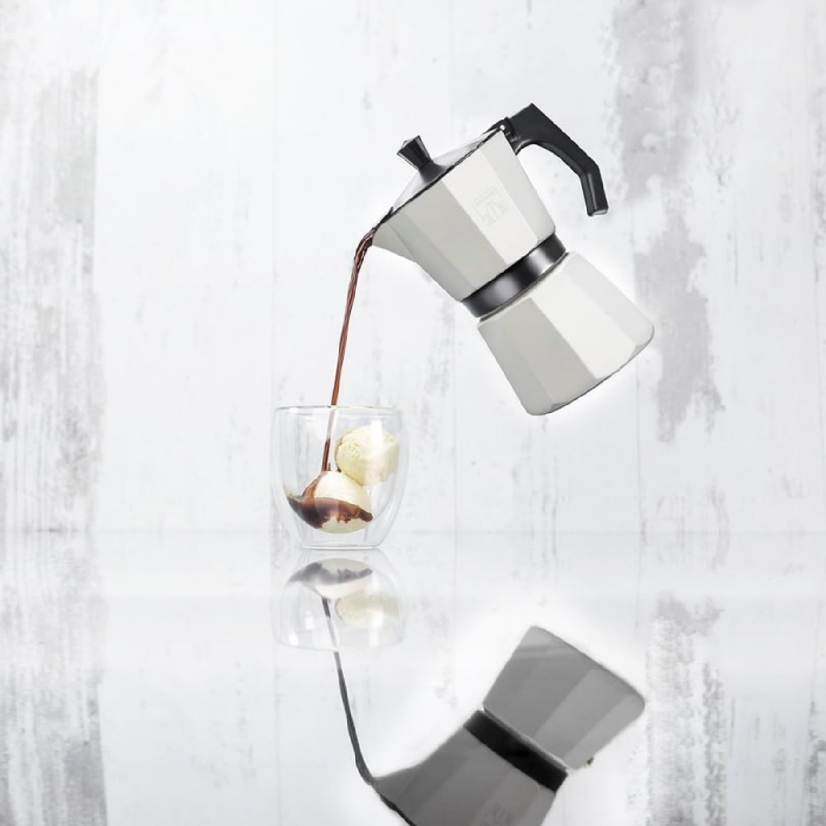 Una caffettiera moka bianca versa caffè in un bicchiere con gelato. Lo sfondo è una parete di legno bianca.