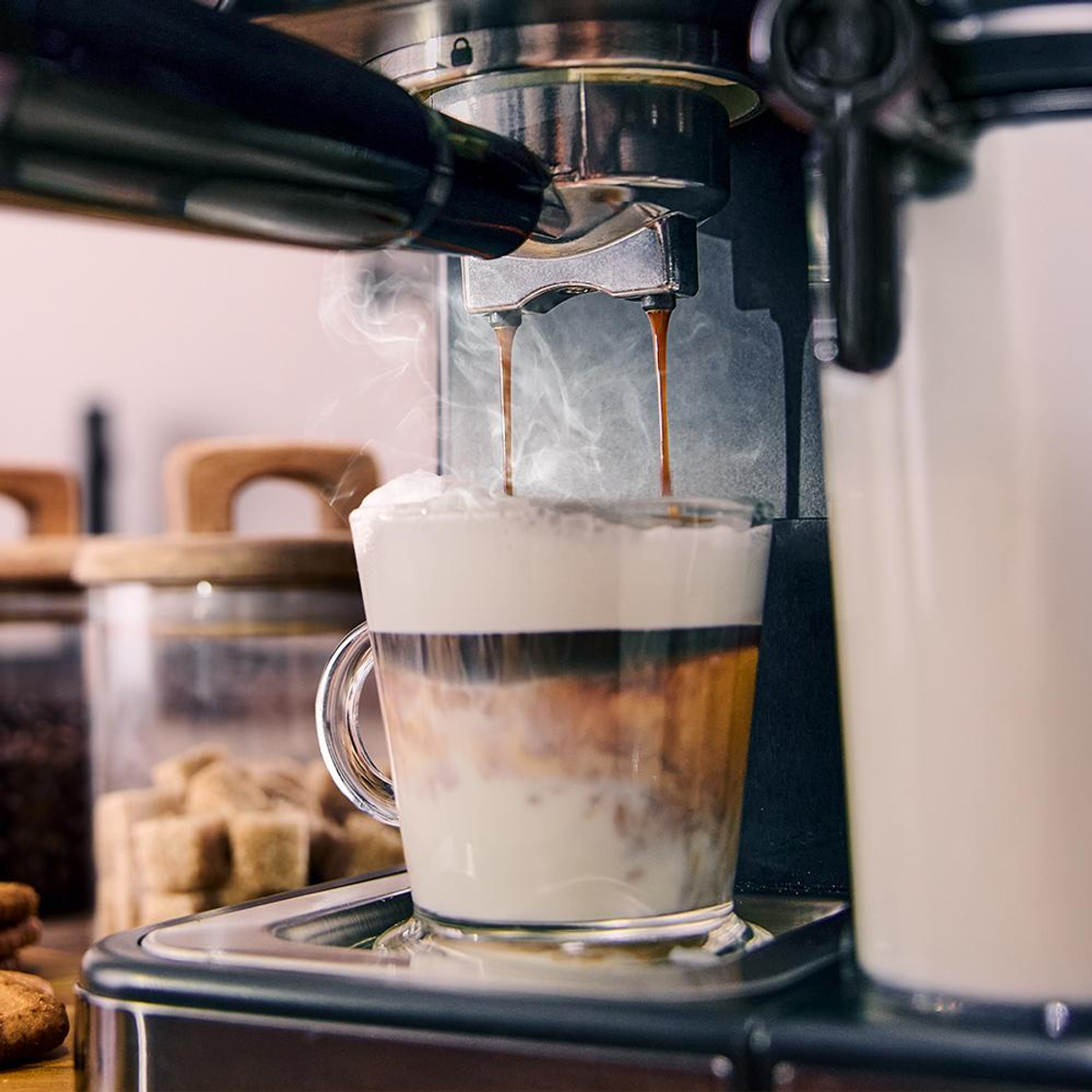 Macchina per espresso che versa caffè in un bicchiere a strati con latte e schiuma. Sale vapore. Sfondo: barattoli e biscotti.