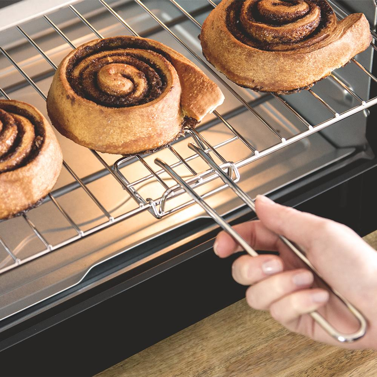 Trzy bułeczki cynamonowe na ruszcie w piekarniku. Ręka trzyma szczypce, aby je wyjąć. Bułeczki są złoto-brązowe z ciemnym wirkiem.